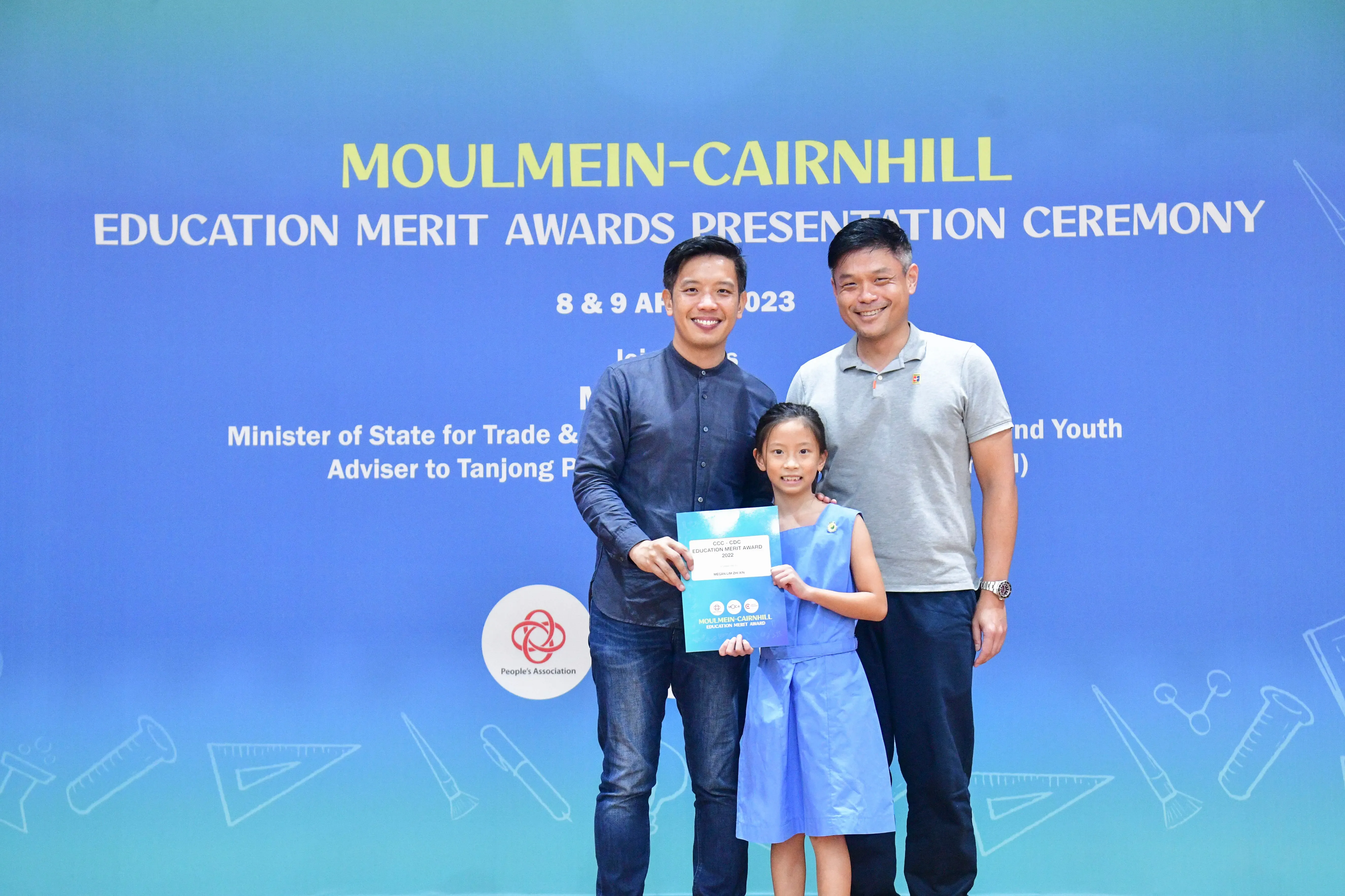 Two men and a young girl hold a certificate in front of a Moulmein-Cairnhill Education Merit Awards backdrop.