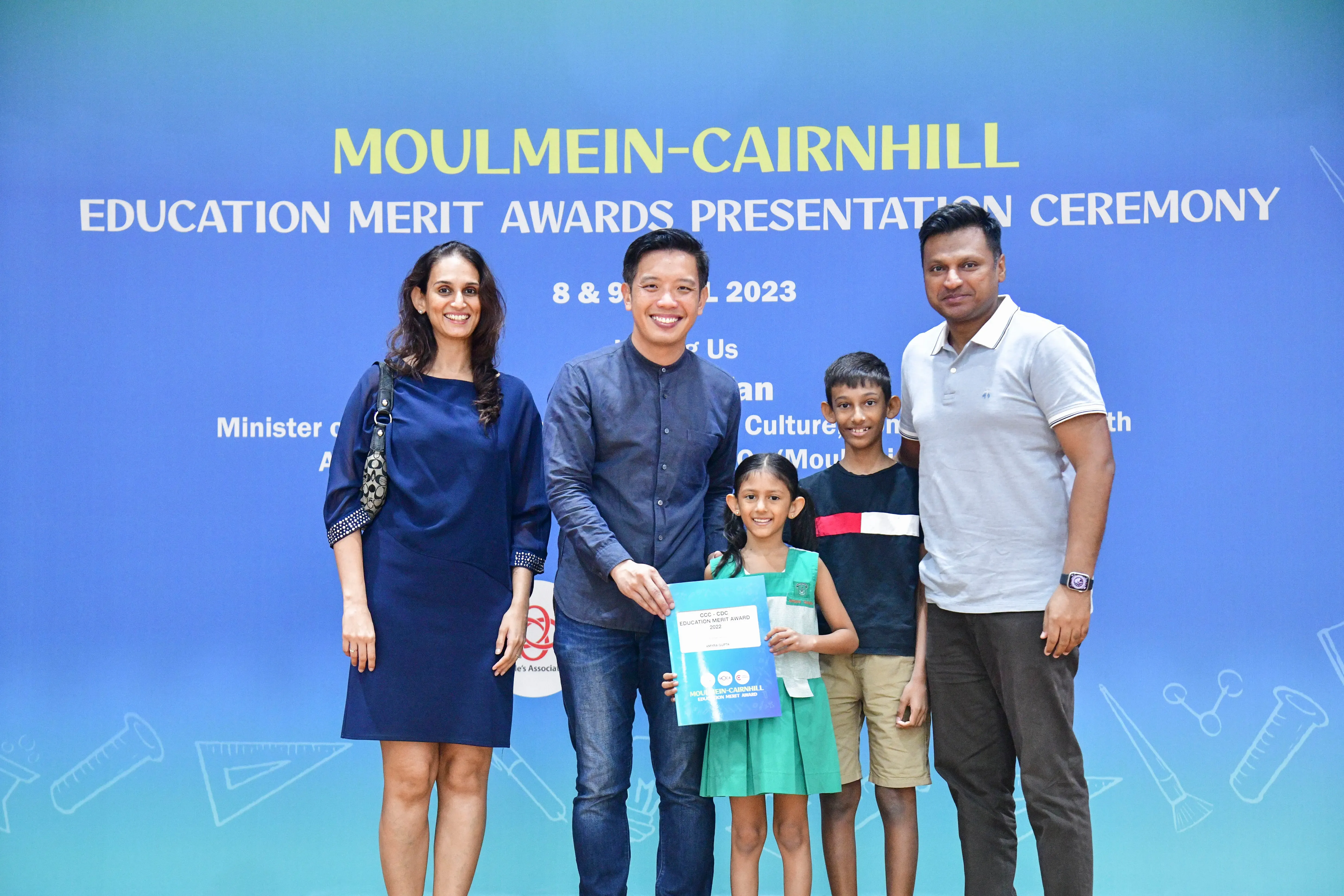Five people pose before a Moulmein-Cairnhill Education Merit Awards Presentation Ceremony banner in 2023.
