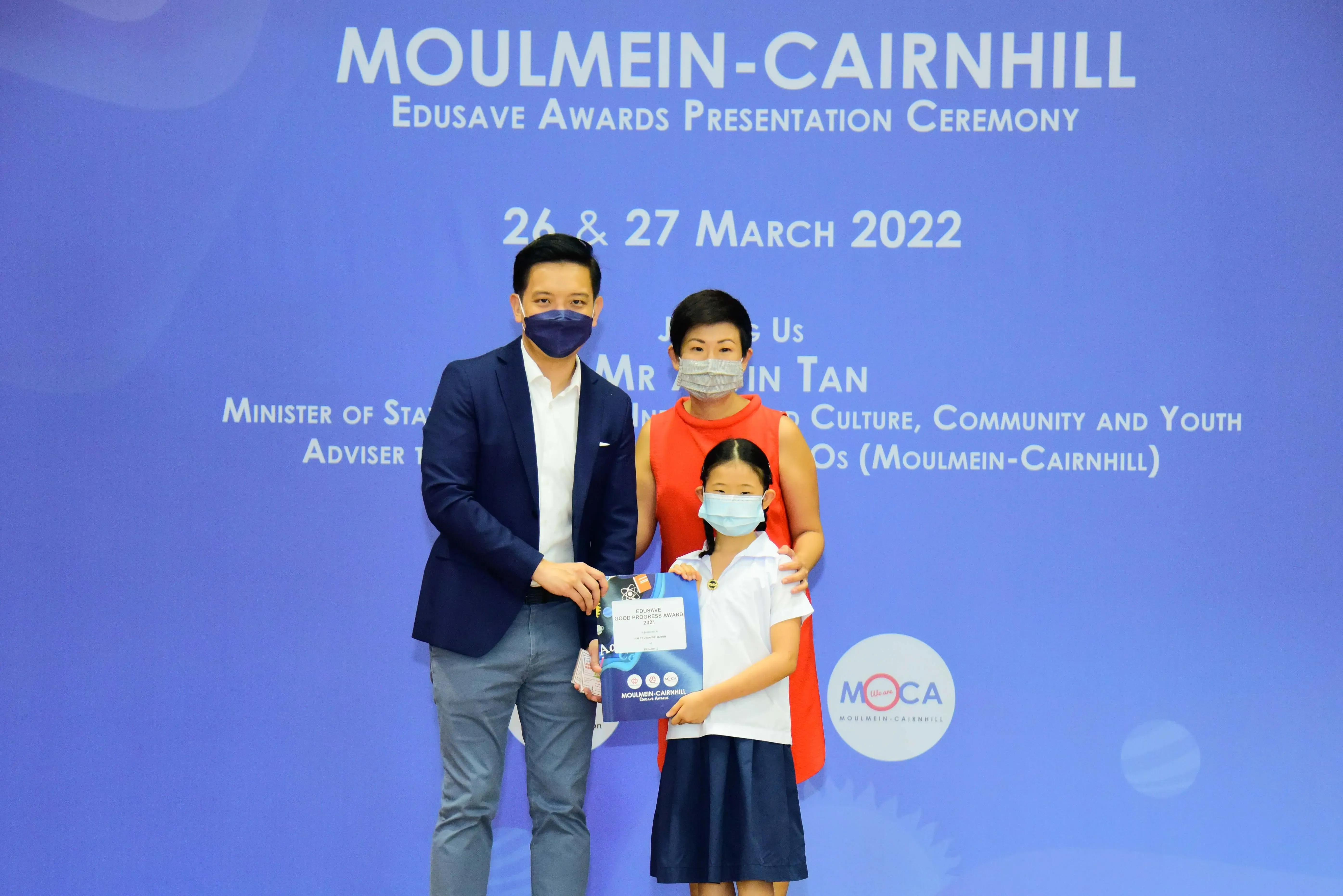 Two masked adults and a masked child holding an award certificate at the Moulmein-Cairnhill Edusave Awards Ceremony 2022.