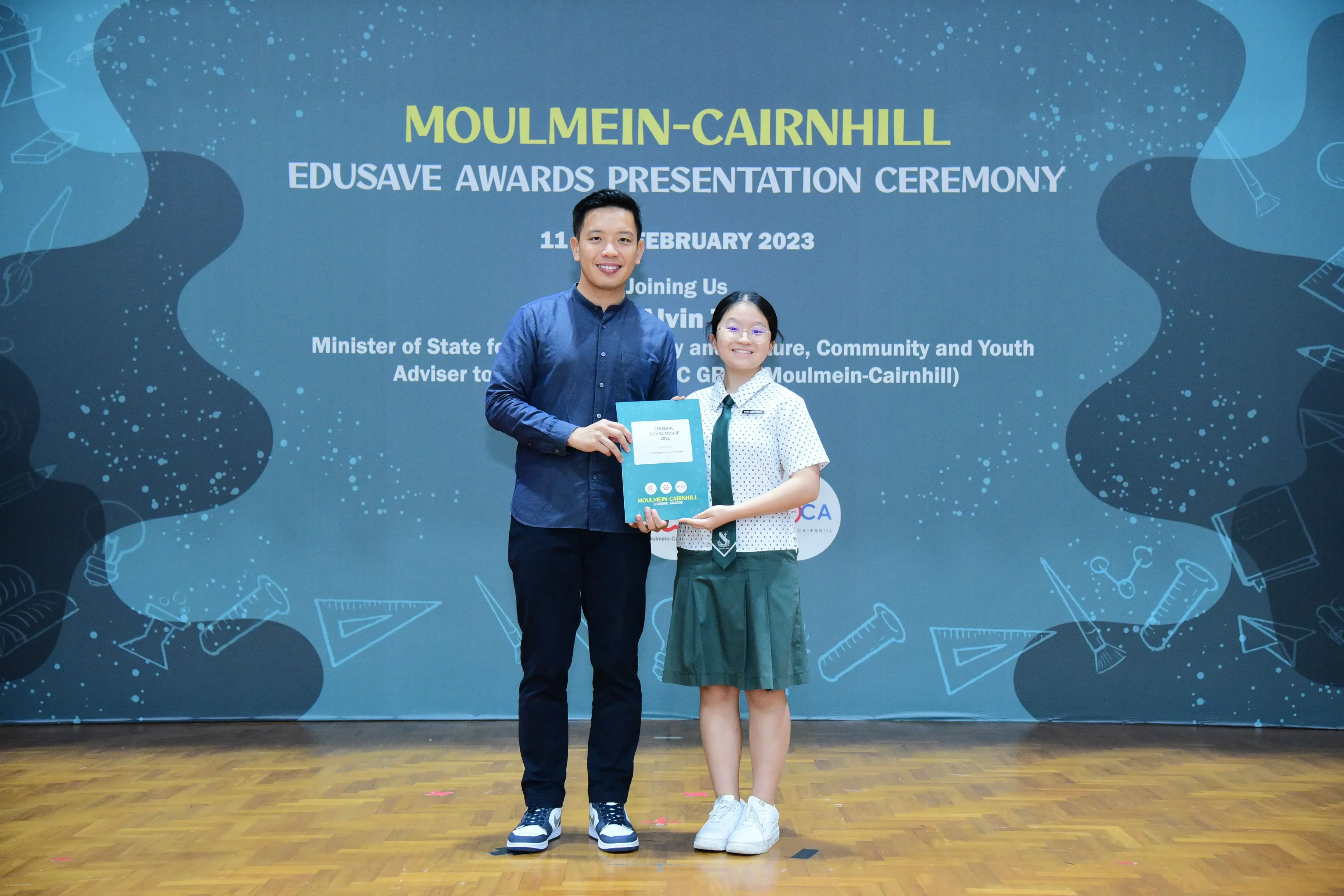 A man and a young girl in school uniform hold a certificate at the Moulmein-Cairnhill Edusave Awards Ceremony 2023.