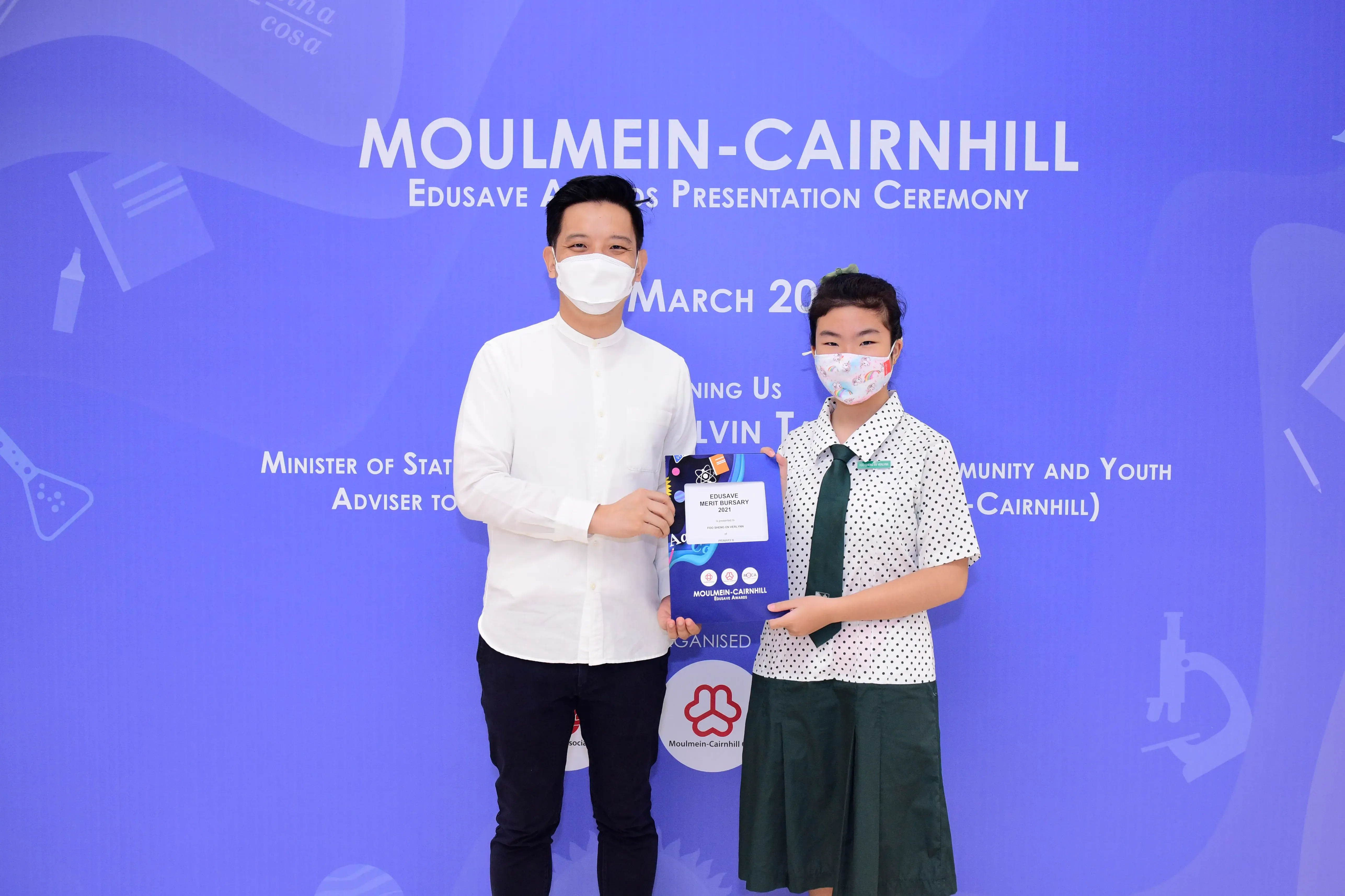Two masked people hold a certificate at the Moulmein-Cairnhill Edusave Awards Presentation Ceremony backdrop.