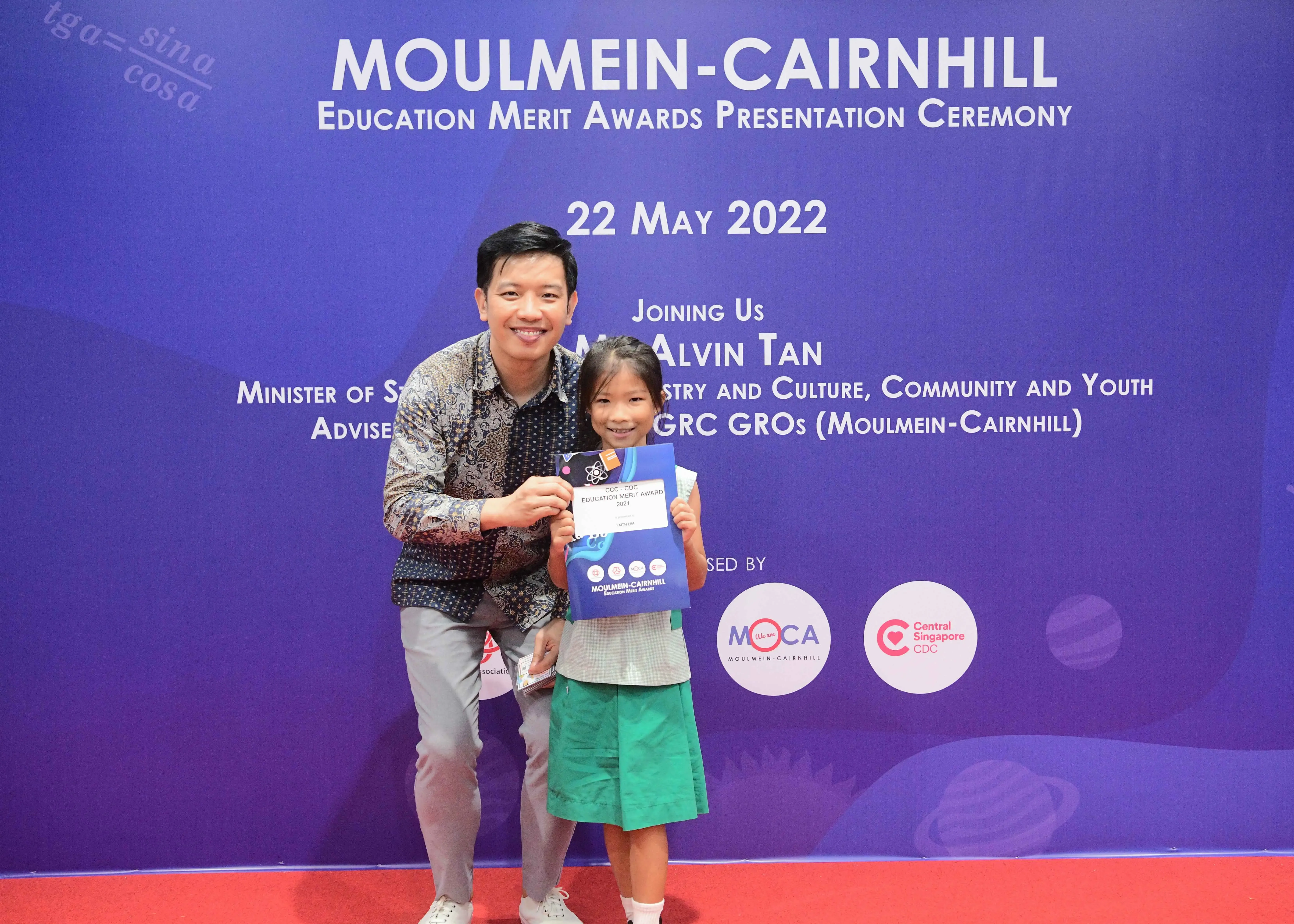 A man and a young girl hold an award certificate in front of a Moulmein-Cairnhill Education Merit Awards banner.