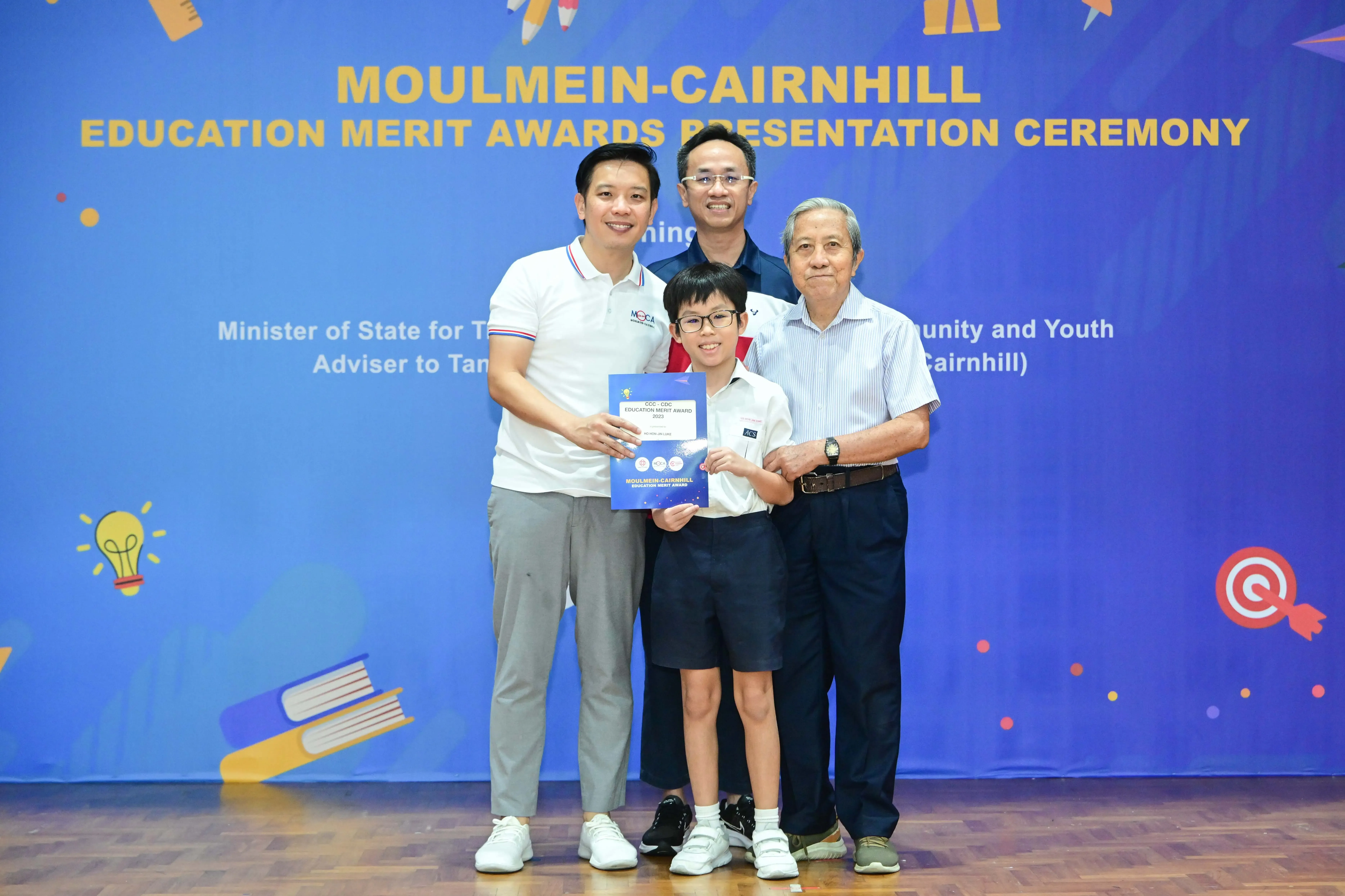 Four people pose at the Moulmein-Cairnhill Education Merit Awards Presentation Ceremony, with a child holding a blue certificate.