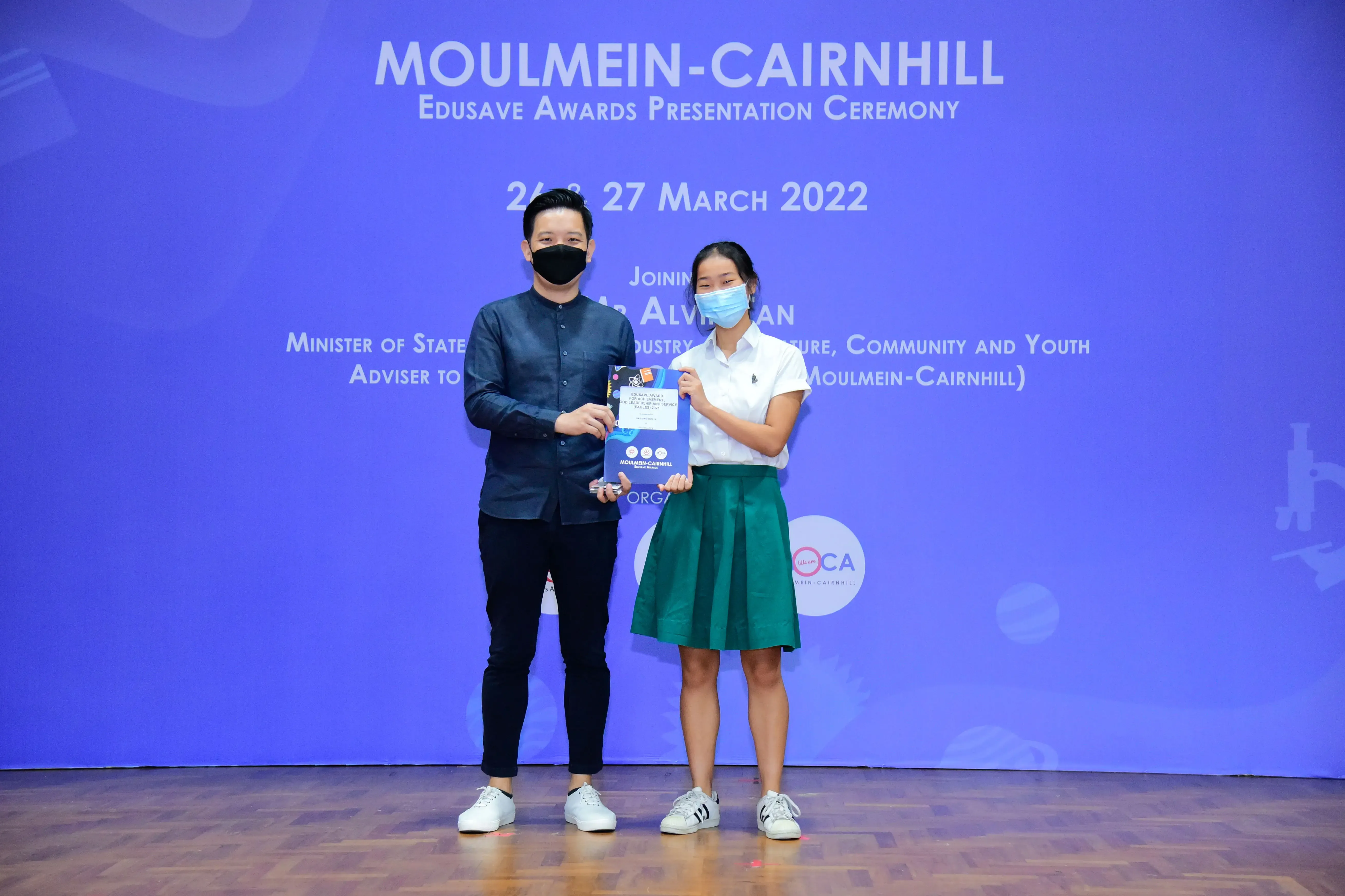 Two masked people hold an award booklet at the Moulmein-Cairnhill Edusave Awards Presentation Ceremony, March 2022.