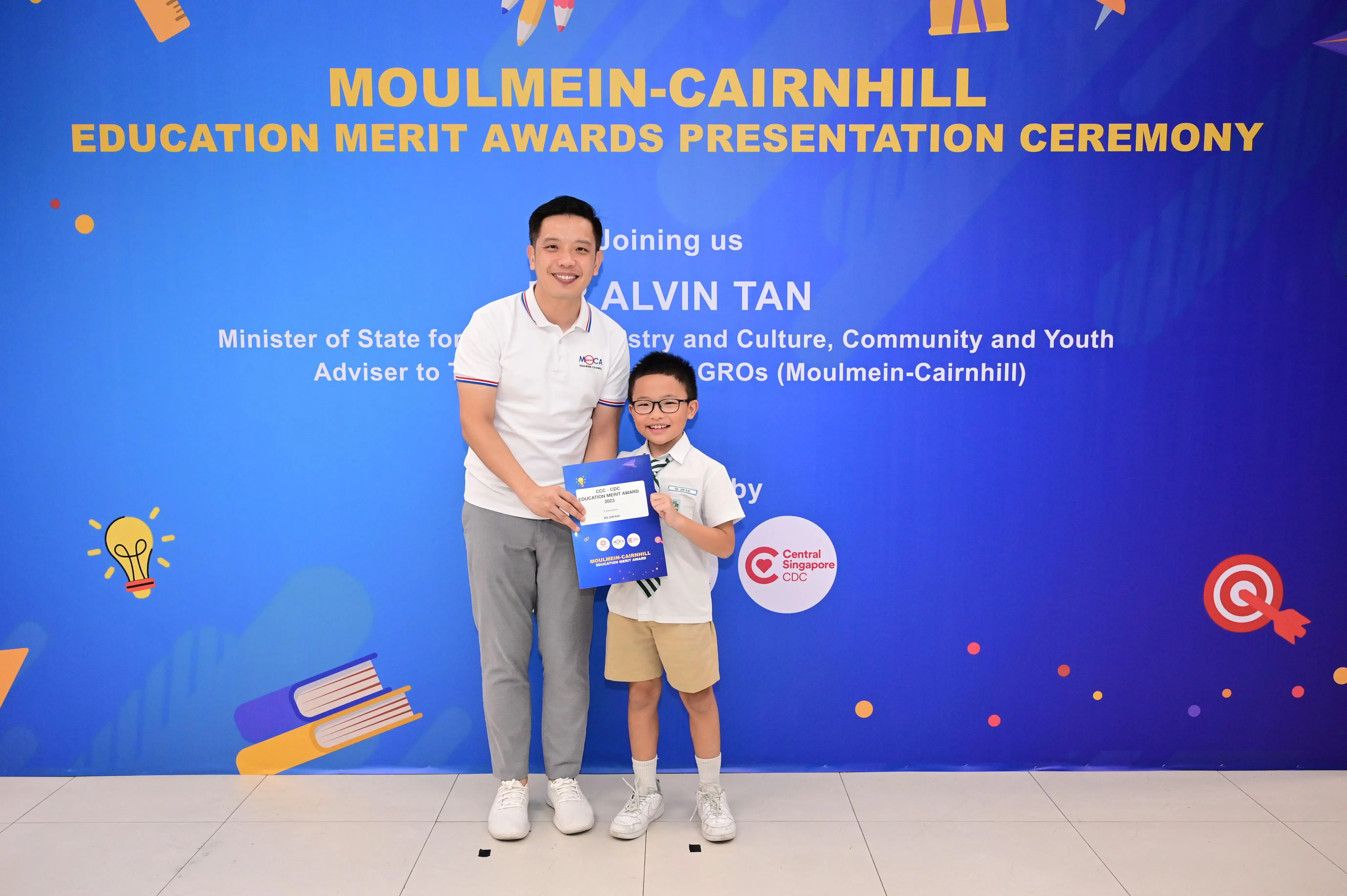 Man and young boy holding a certificate in front of a Moulmein-Cairnhill Education Merit Awards banner.