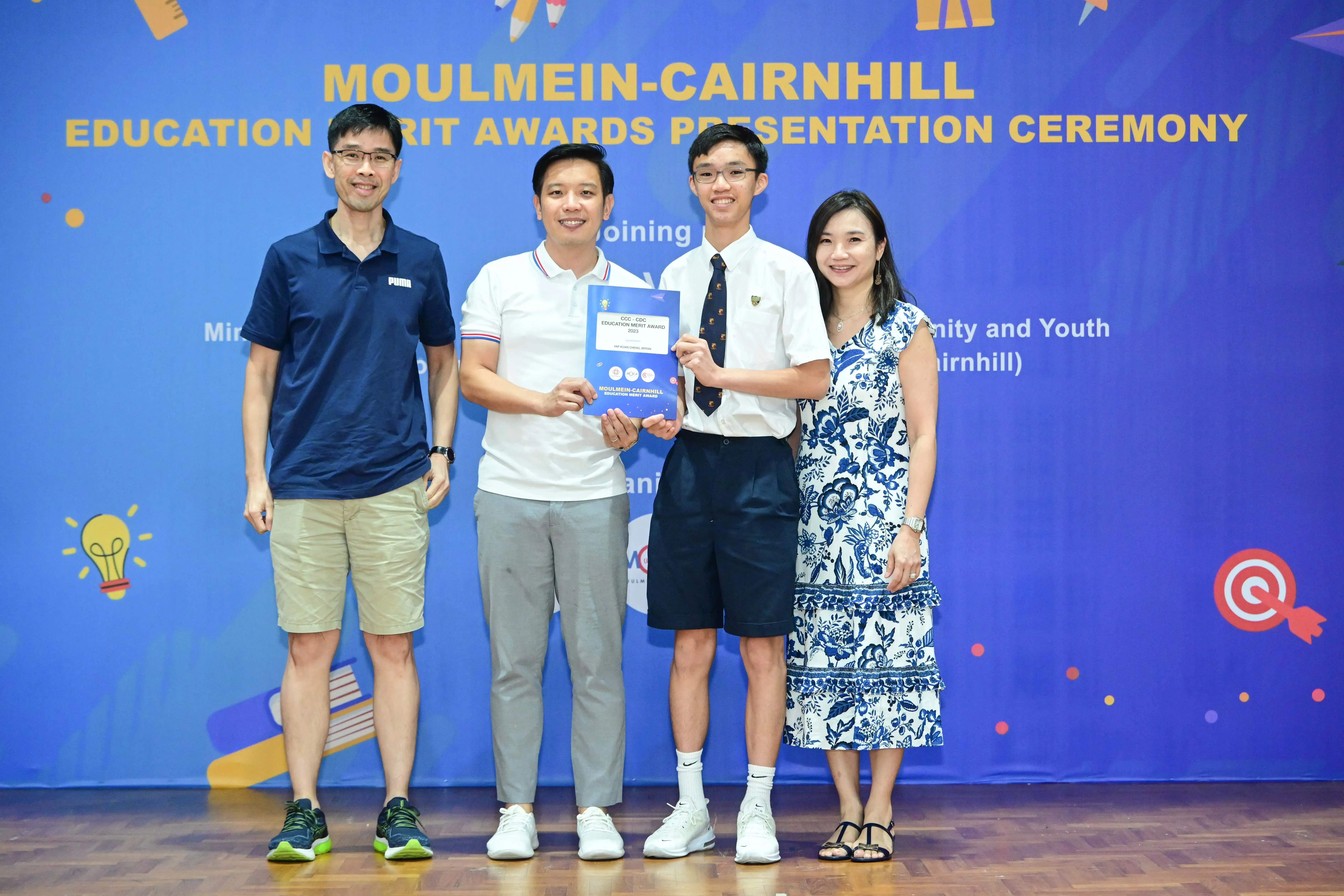 Four people pose at the Moulmein-Cairnhill Education Merit Awards Presentation Ceremony, with two holding a blue award certificate.