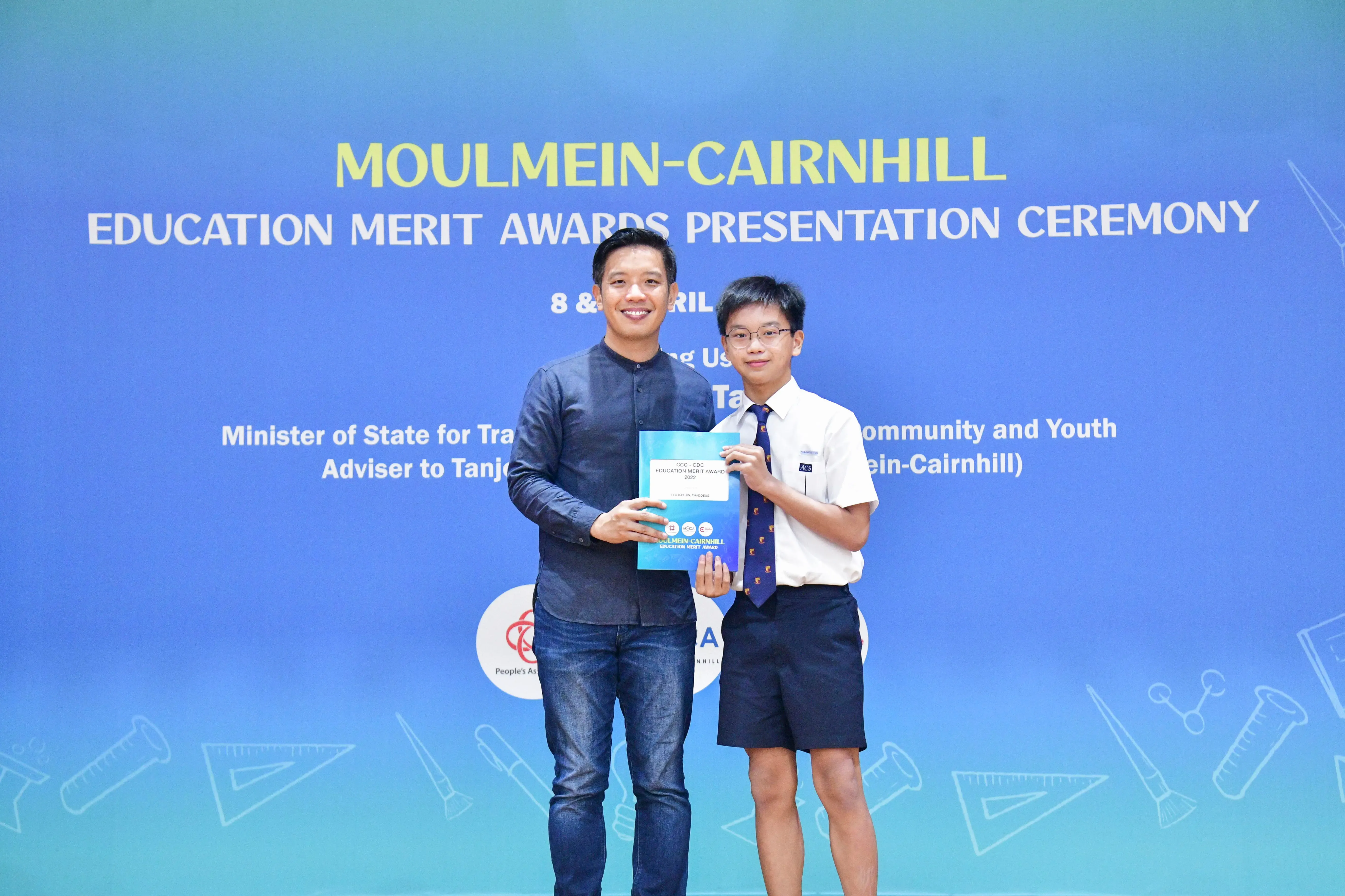 Two people hold a certificate together in front of a Moulmein-Cairnhill Education Merit Awards Presentation Ceremony banner.