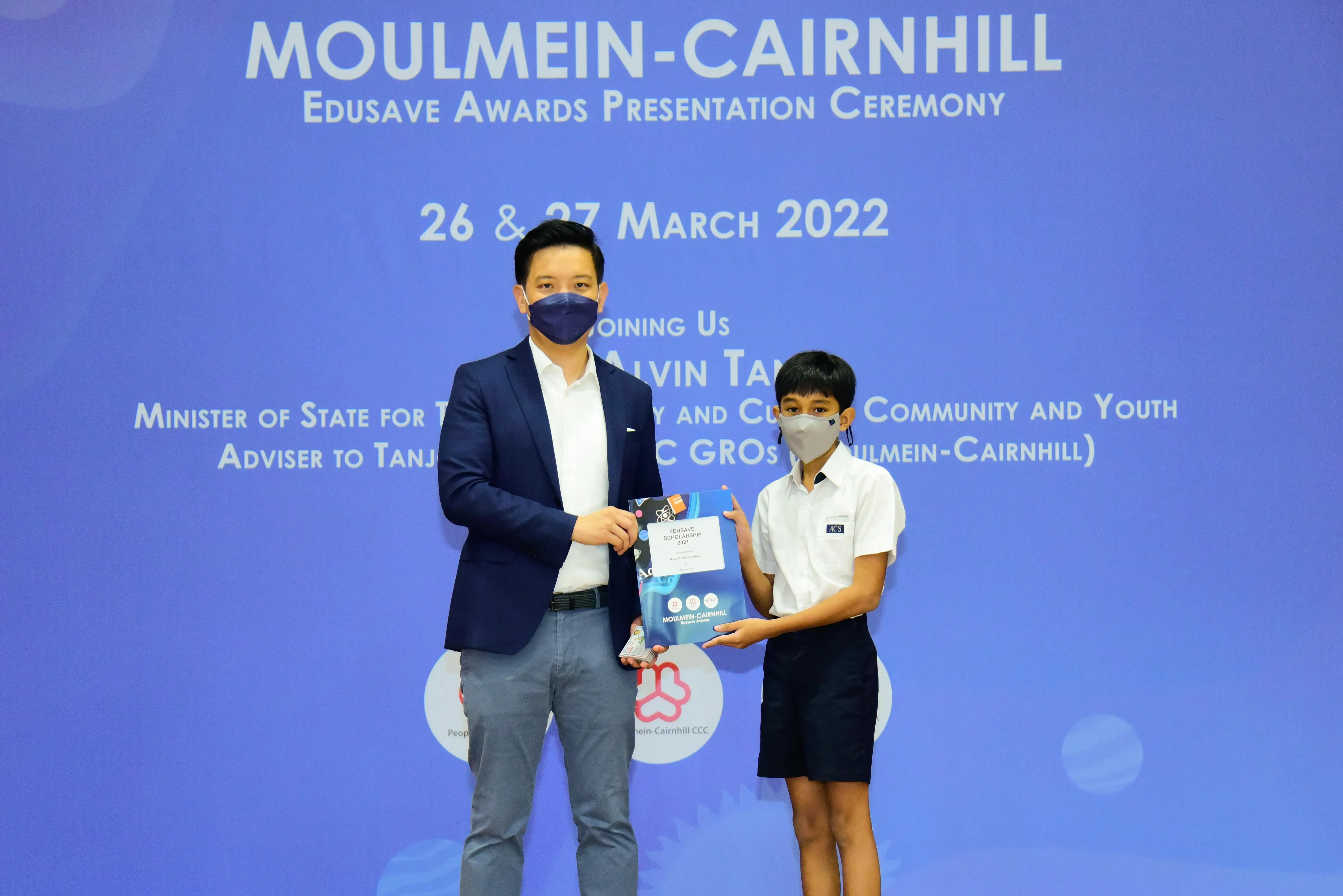 An adult and a masked student hold an Edusave Scholarship certificate at the Moulmein-Cairnhill Awards Ceremony, March 2022.
