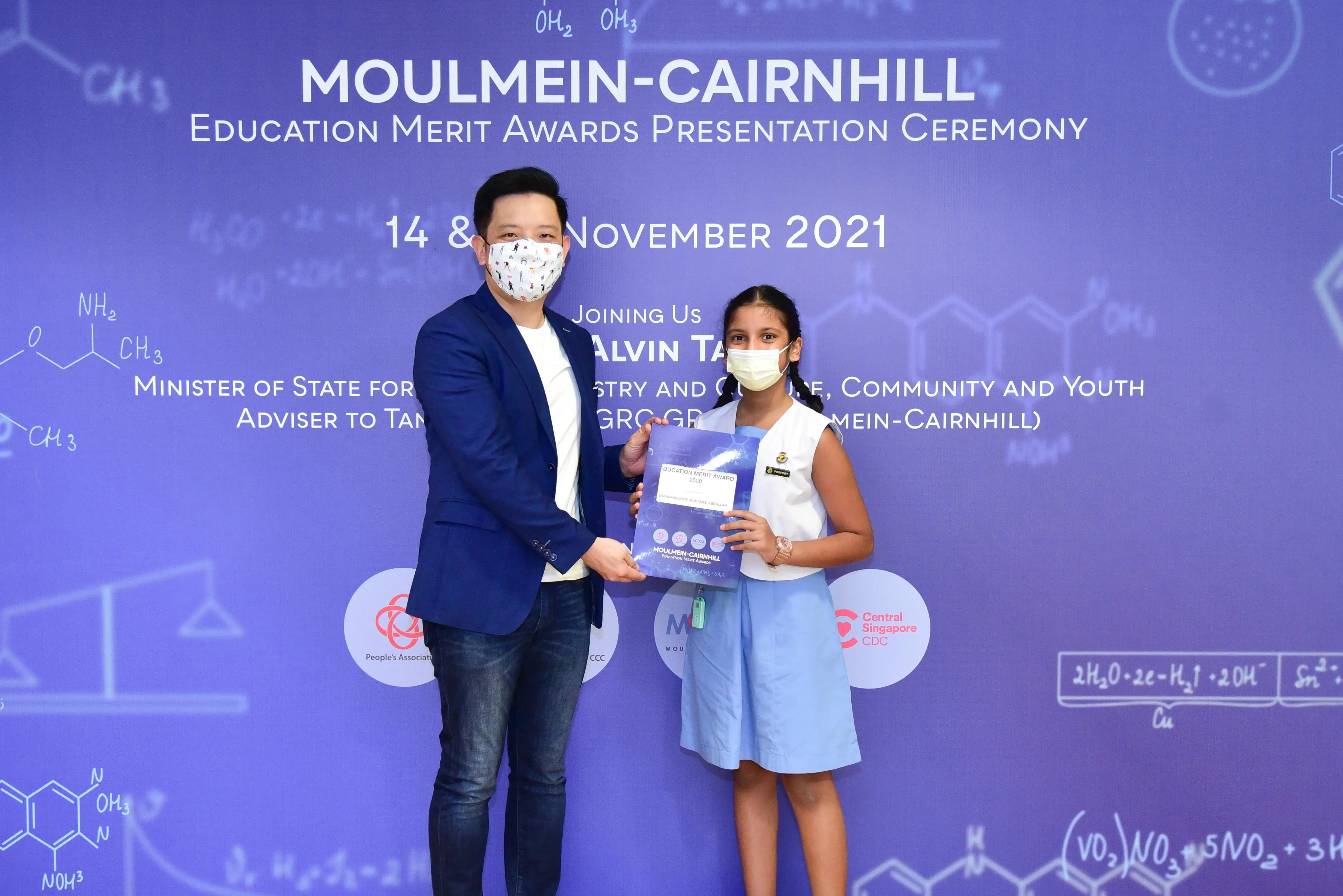 A masked man presents an award certificate to a masked young girl at the Moulmein-Cairnhill Education Merit Awards 2021.