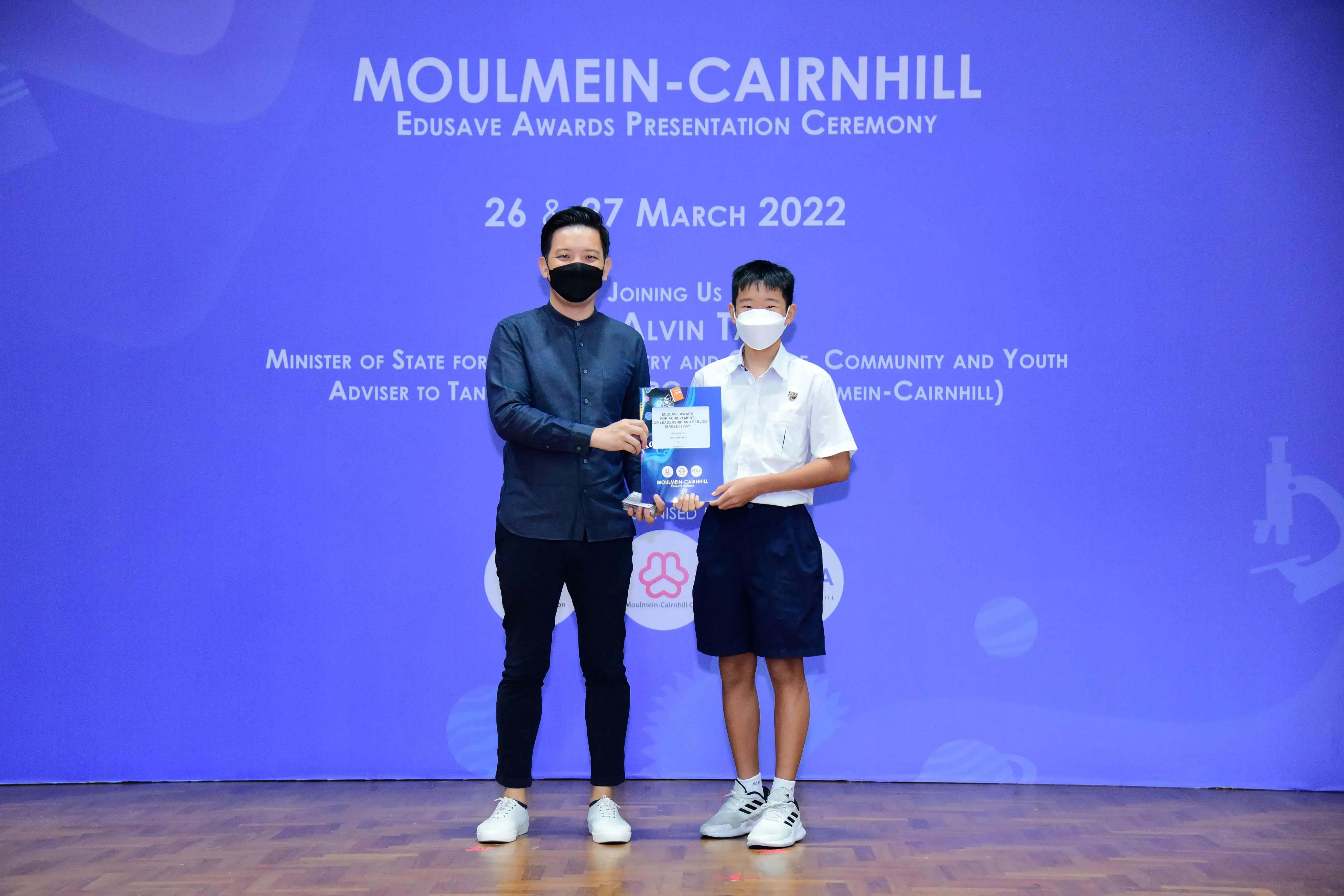 Two masked individuals hold an award at the Moulmein-Cairnhill Edusave Awards Presentation Ceremony, March 2022.