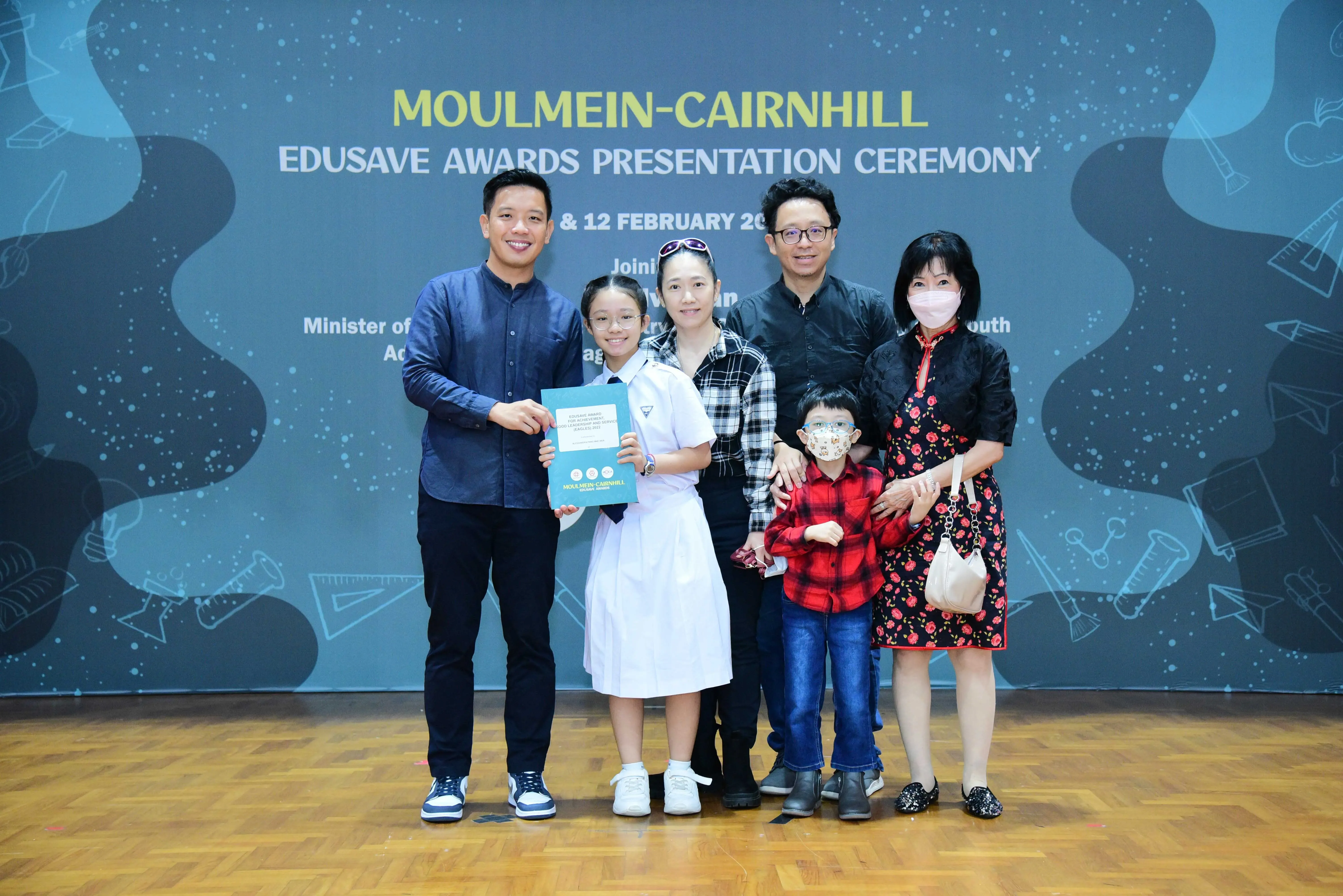 Five adults and two children pose at the Moulmein-Cairnhill Edusave Awards Presentation Ceremony, holding a certificate.