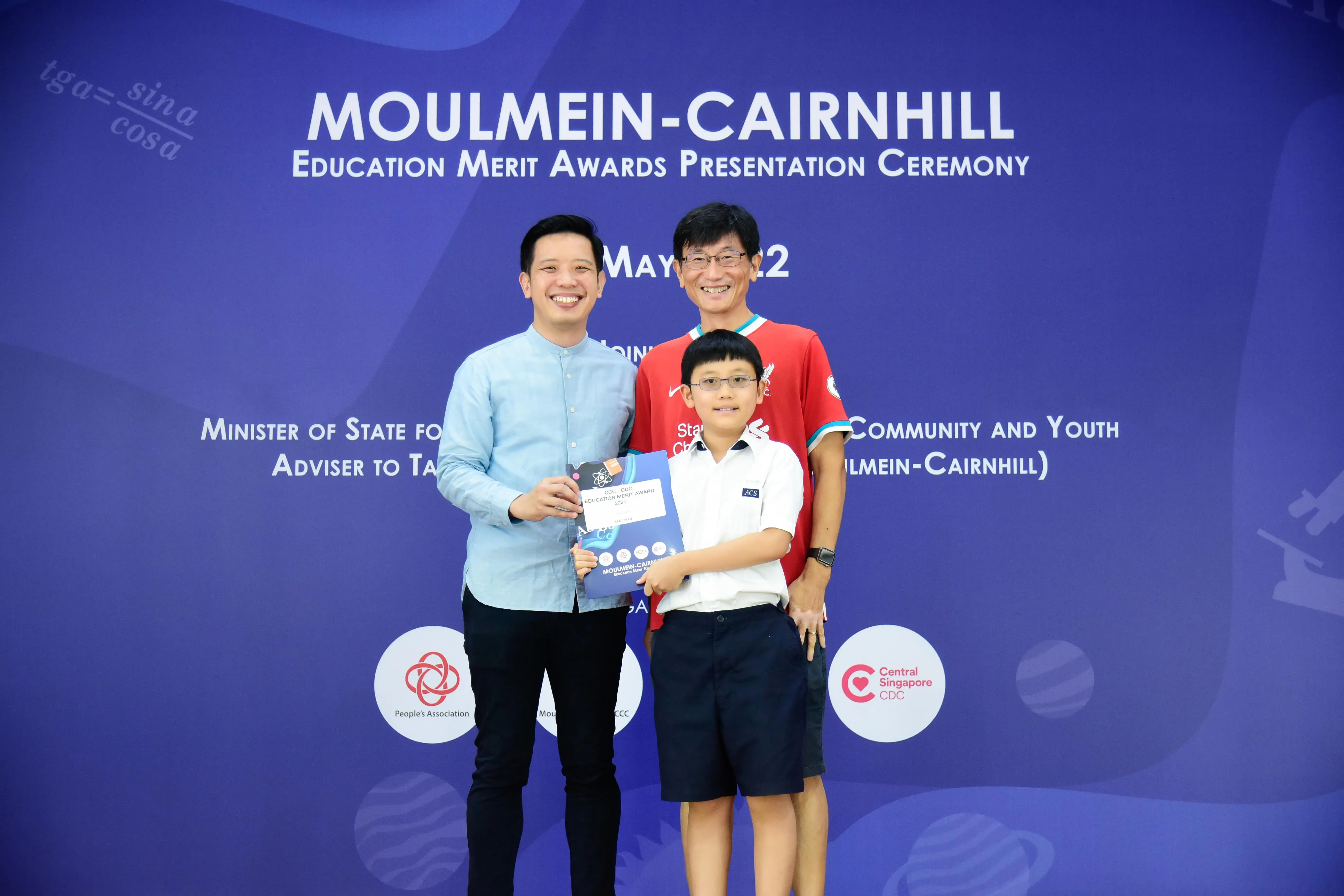 Two men and a boy holding an award certificate pose at the Moulmein-Cairnhill Education Merit Awards ceremony.