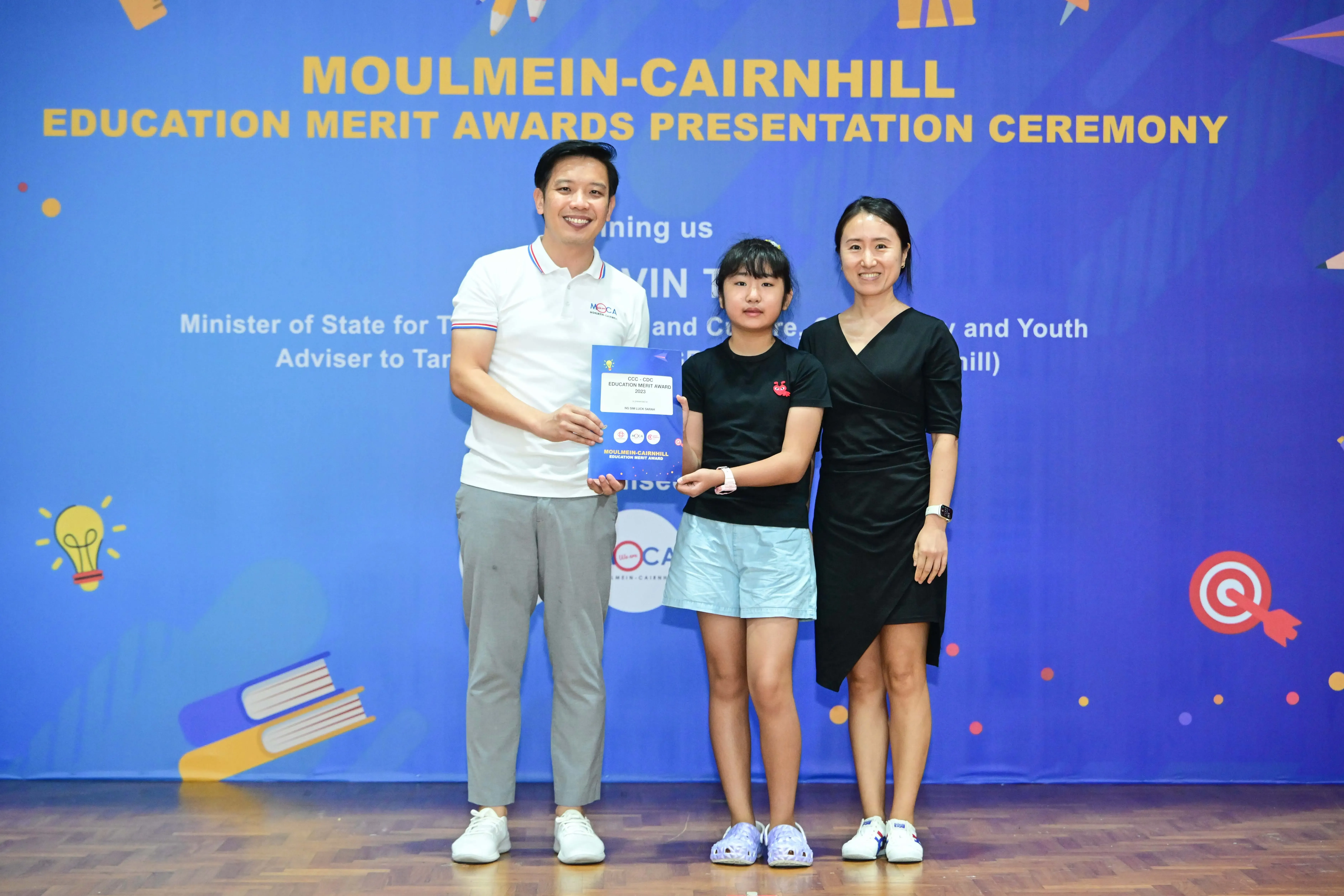 Three people pose holding a certificate at the Moulmein-Cairnhill Education Merit Awards Presentation Ceremony.