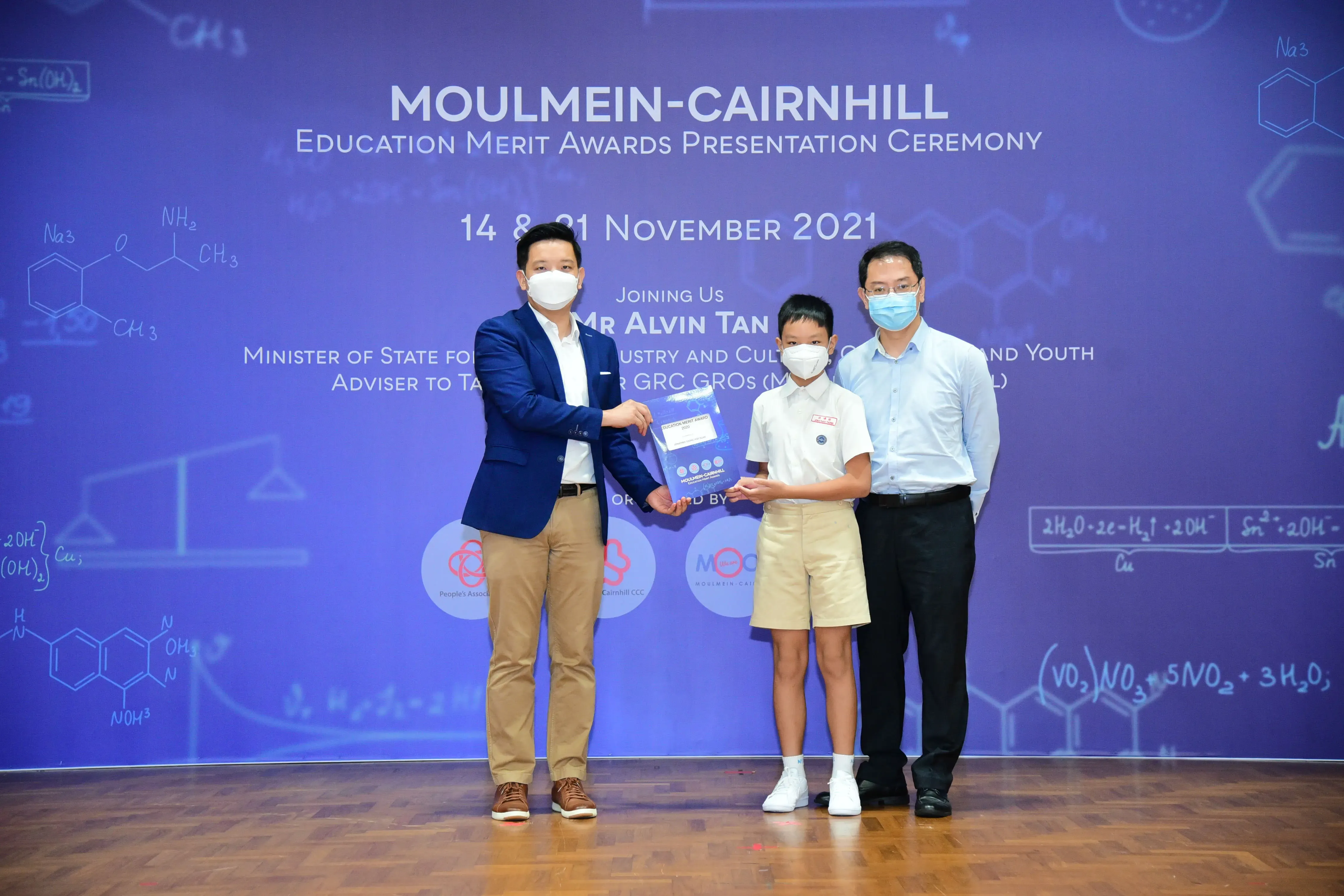 Two masked adults present an award to a masked child at the Moulmein-Cairnhill Education Merit Awards Ceremony 2021.