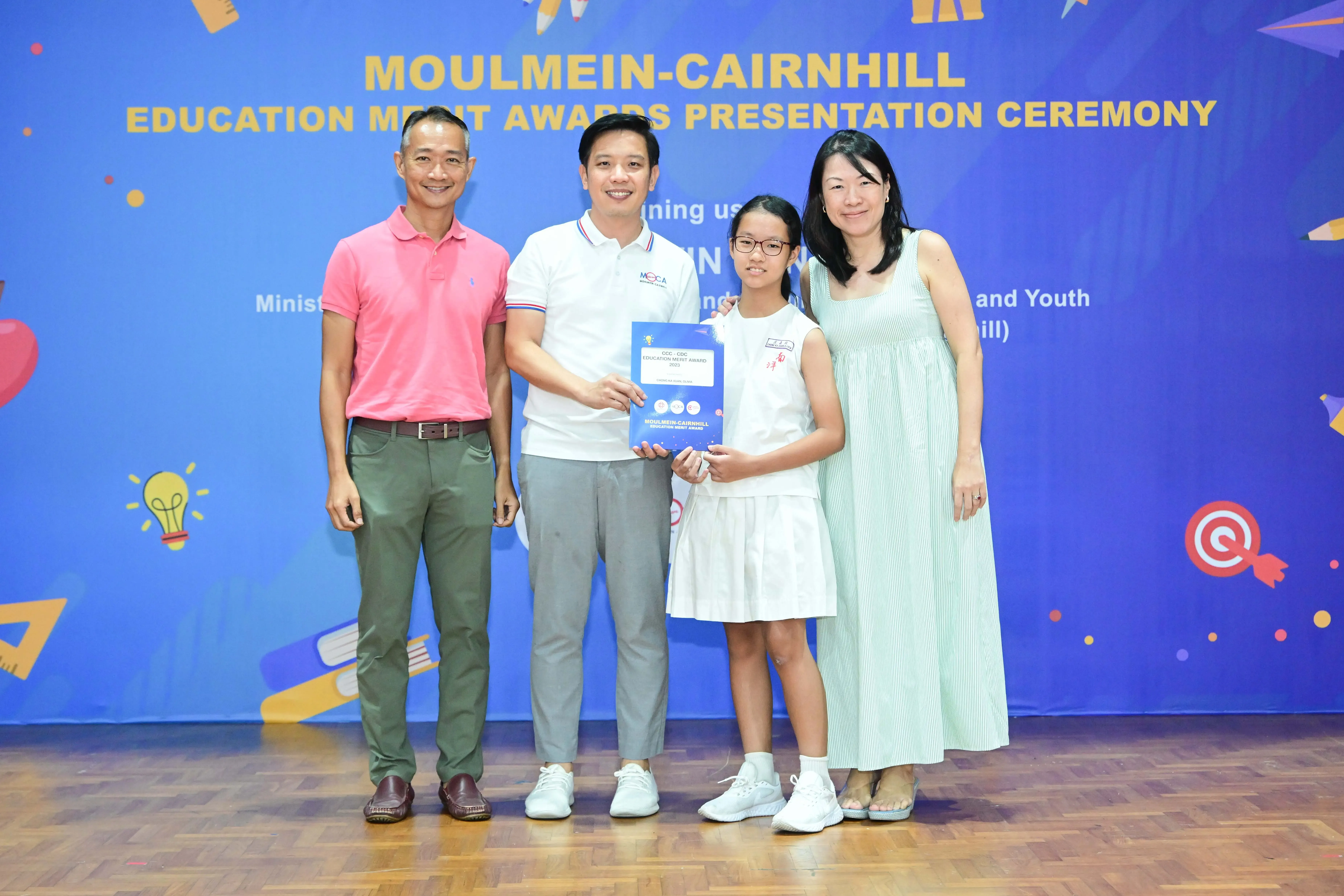 Four people pose at Moulmein-Cairnhill Education Merit Awards ceremony, with a girl holding an award certificate.