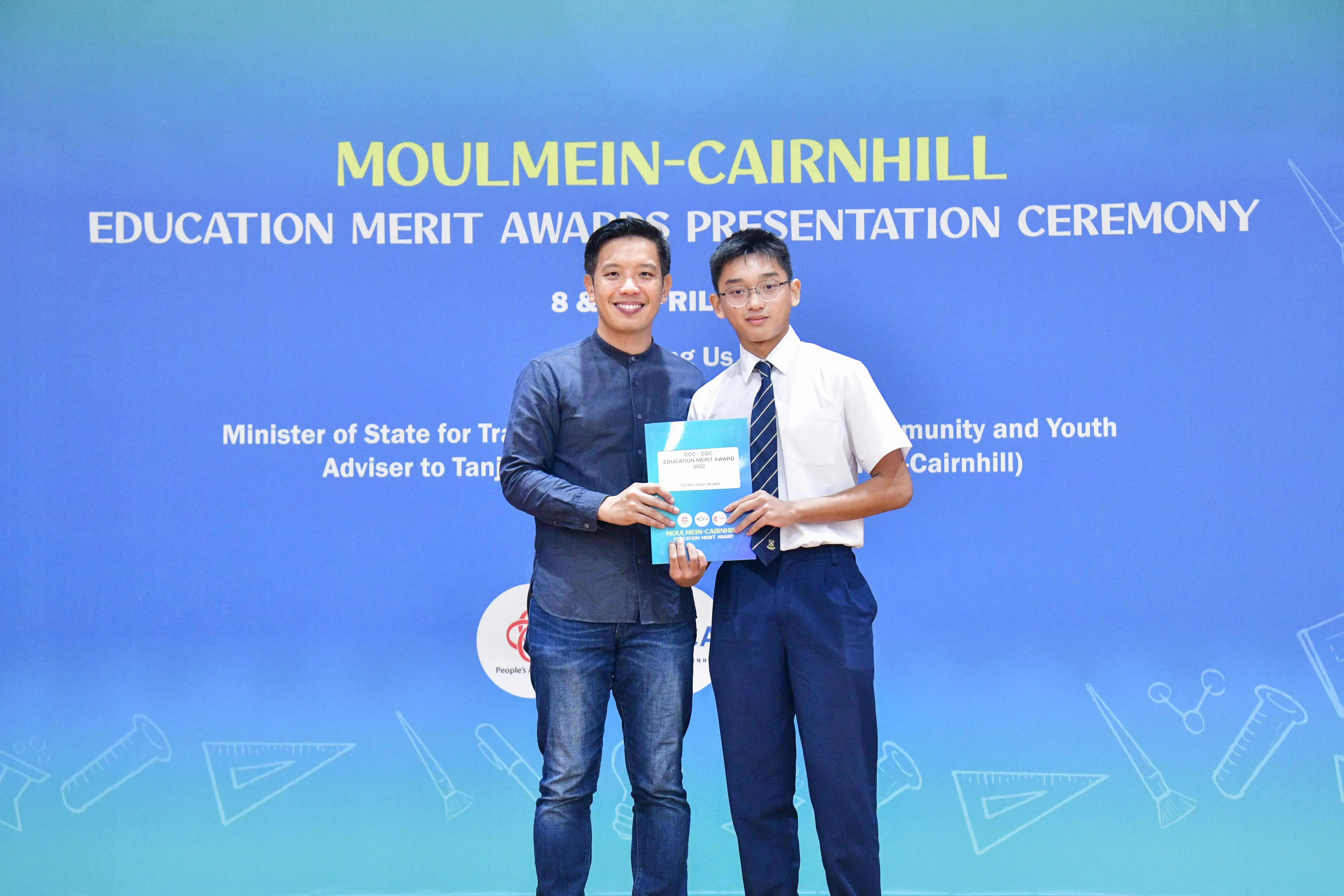 Two males hold a certificate at the Moulmein-Cairnhill Education Merit Awards Presentation Ceremony backdrop.