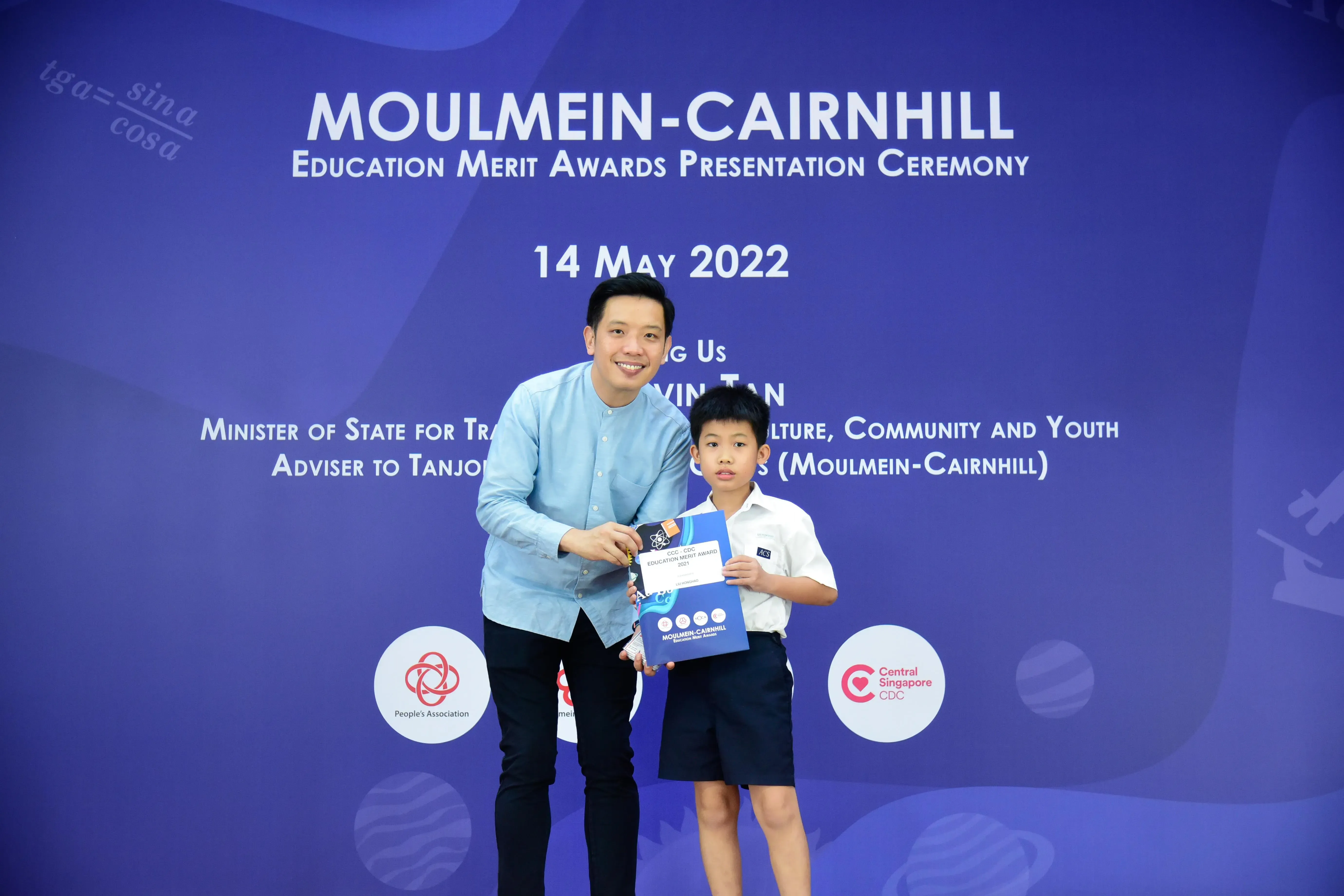 A man and a young boy hold an award certificate before a Moulmein-Cairnhill Education Merit Awards backdrop dated 14 May 2022.