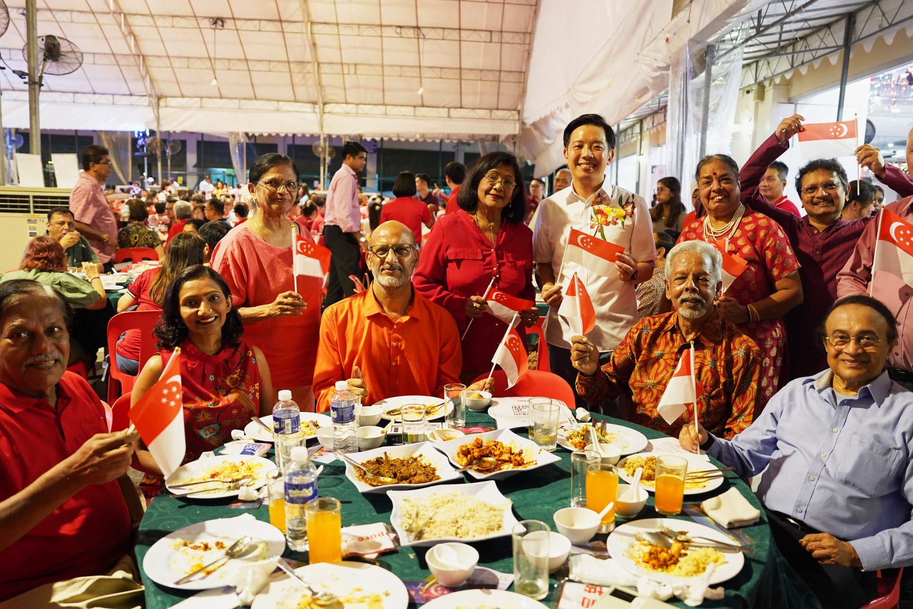 A group of smiling people in red clothing hold Singapore flags around a dinner table filled with food at a festive event.