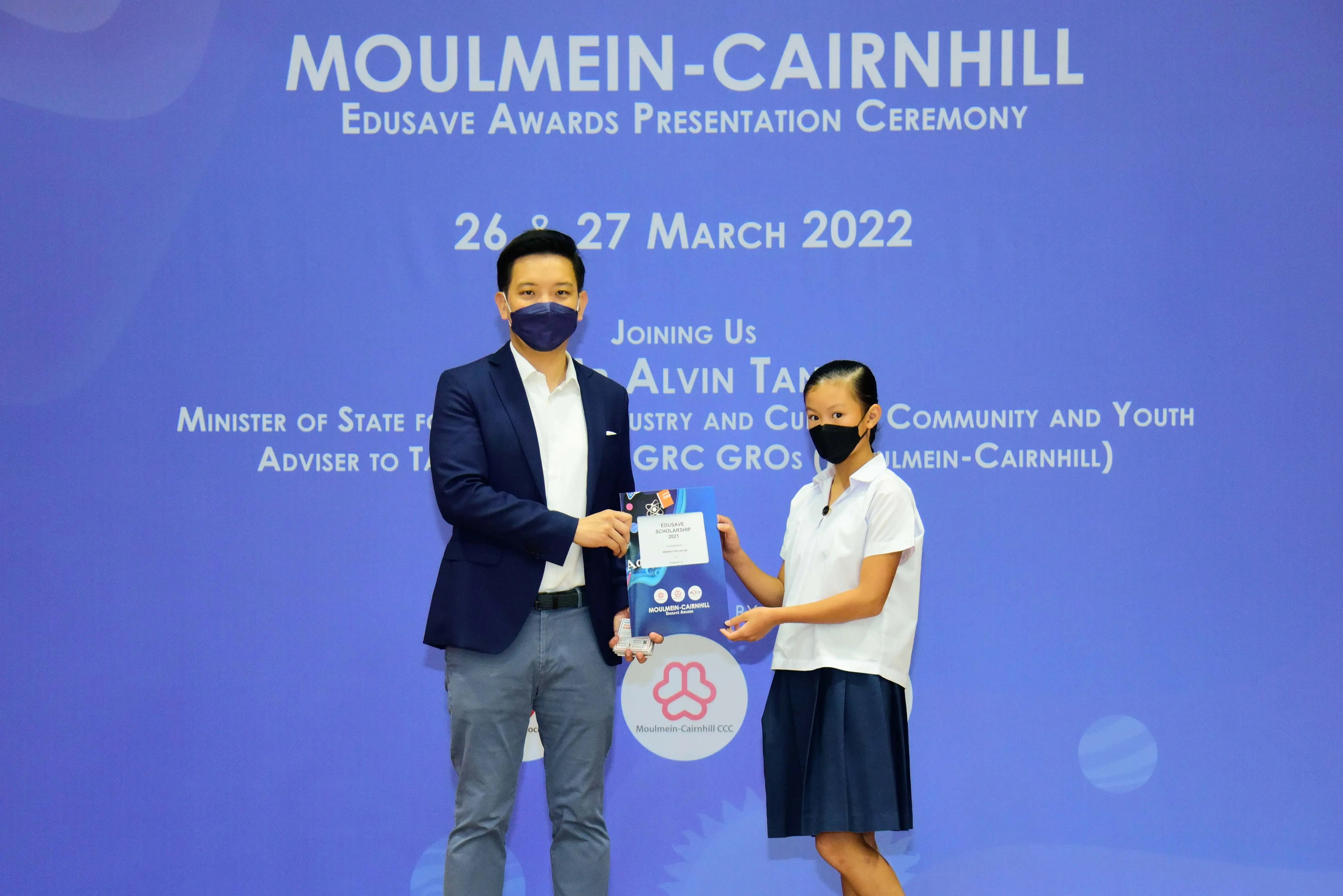 A masked man in a blazer presents an award to a masked young girl at the Moulmein-Cairnhill Edusave ceremony, March 2022.