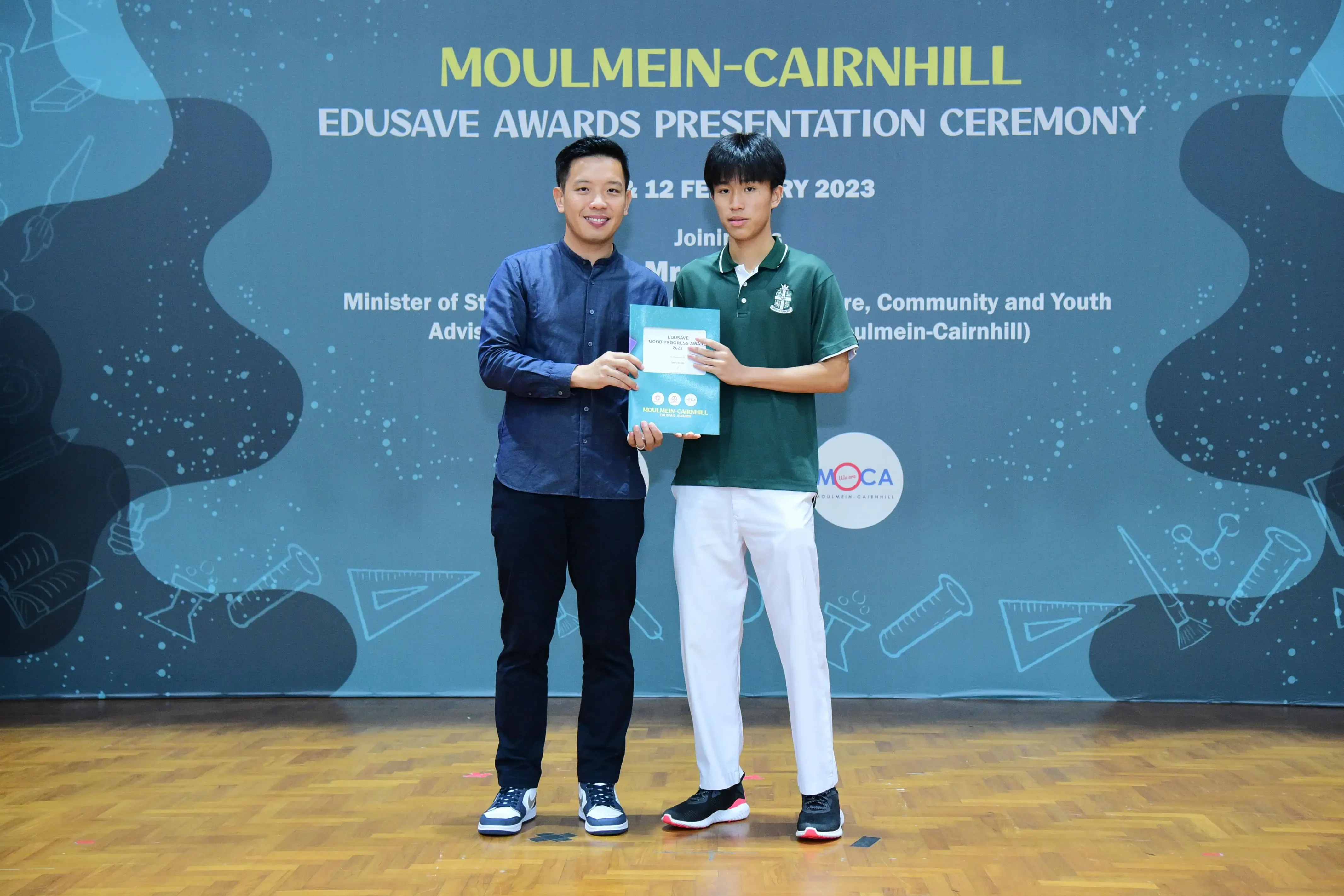 Two people hold a certificate together on stage at the Moulmein-Cairnhill Edusave Awards Presentation Ceremony 2023.