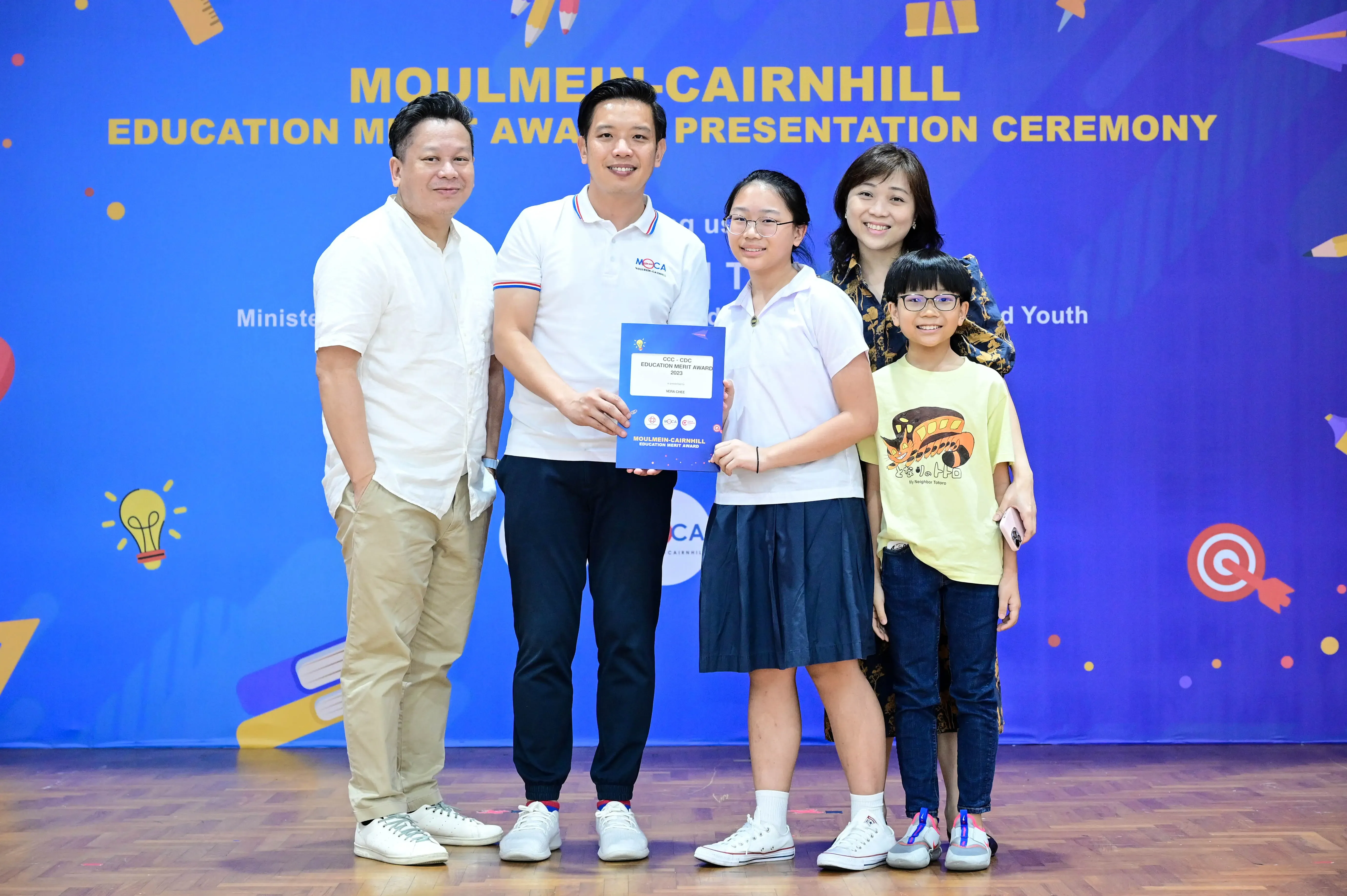 Five people pose at the Moulmein-Cairnhill Education Merit Award Presentation Ceremony, with a student holding a certificate.