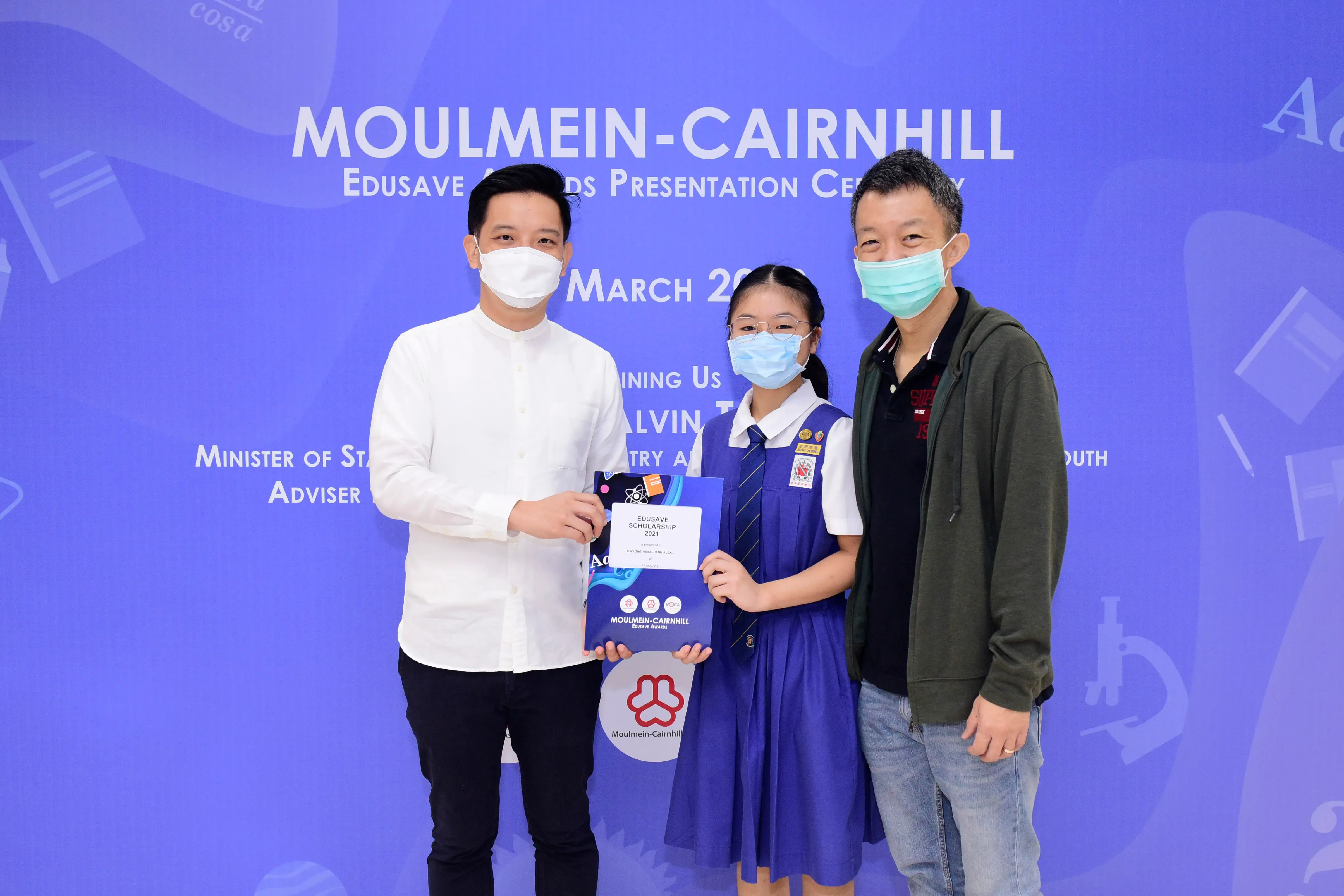 Three masked people pose at the Moulmein-Cairnhill Edusave Awards Presentation Ceremony, with a girl holding a scholarship certificate.