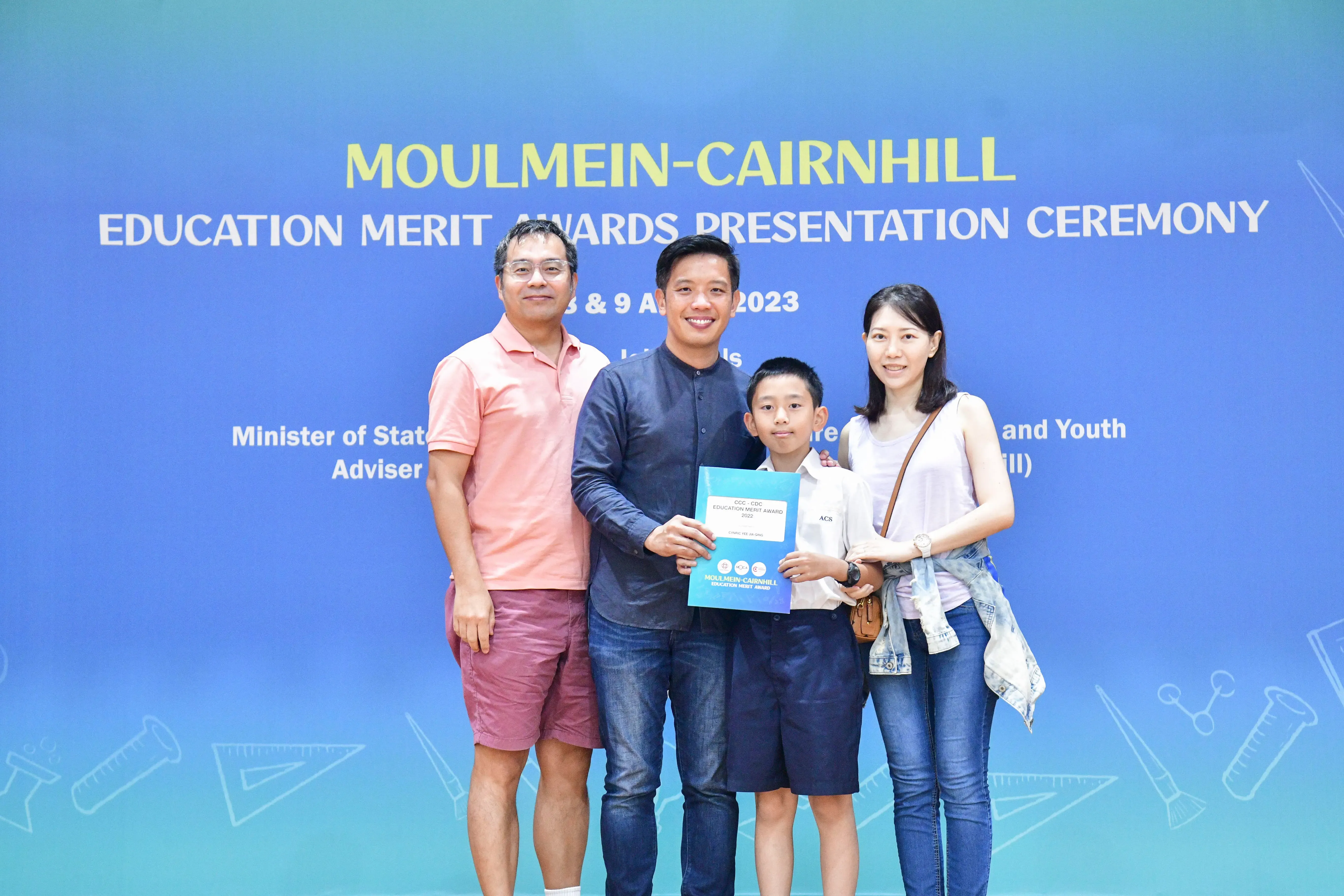 Four people pose in front of a blue Moulmein-Cairnhill Education Merit Awards Presentation Ceremony banner in 2023.