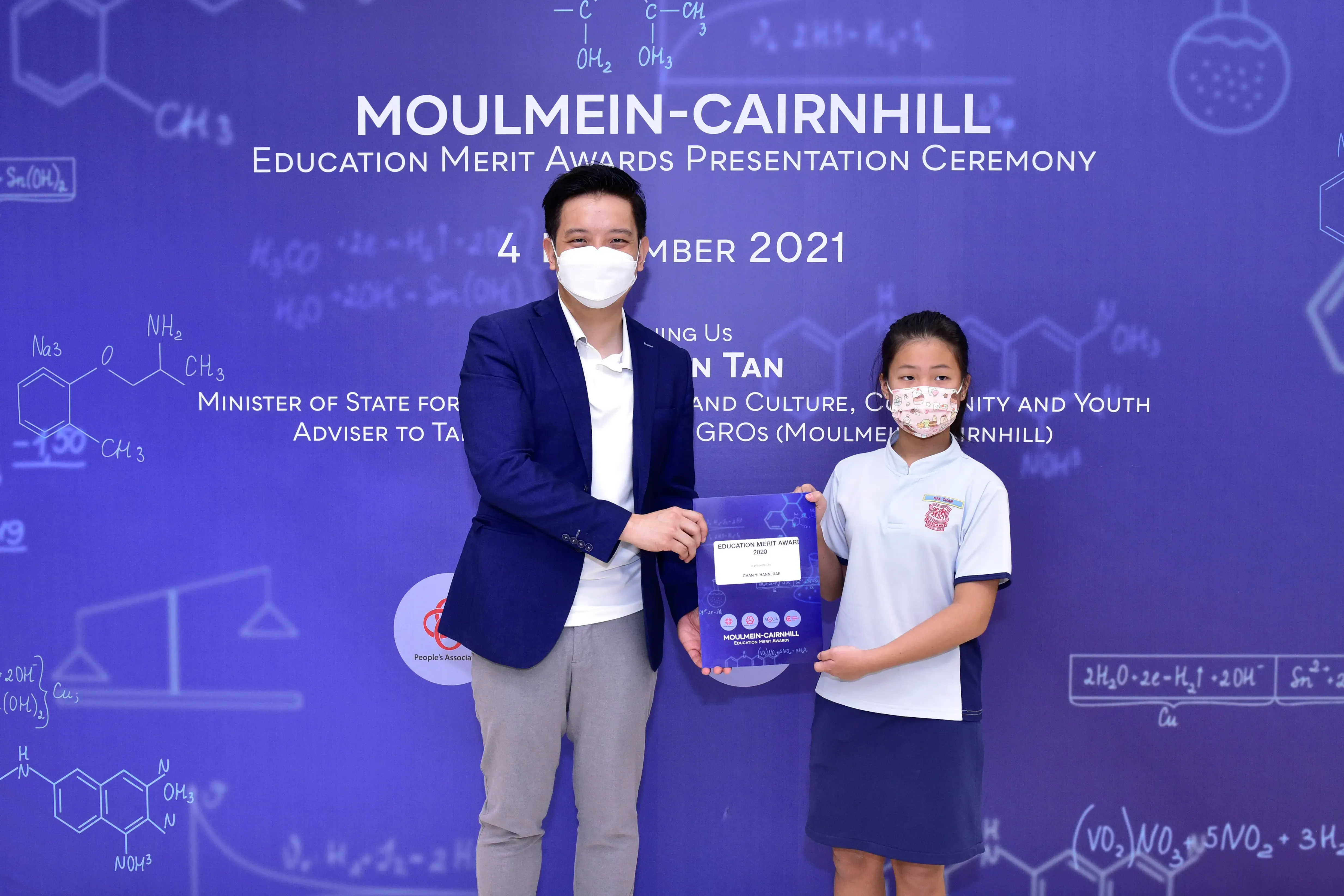 A masked adult and young student hold an award certificate at the Moulmein-Cairnhill Education Merit Awards 2021.