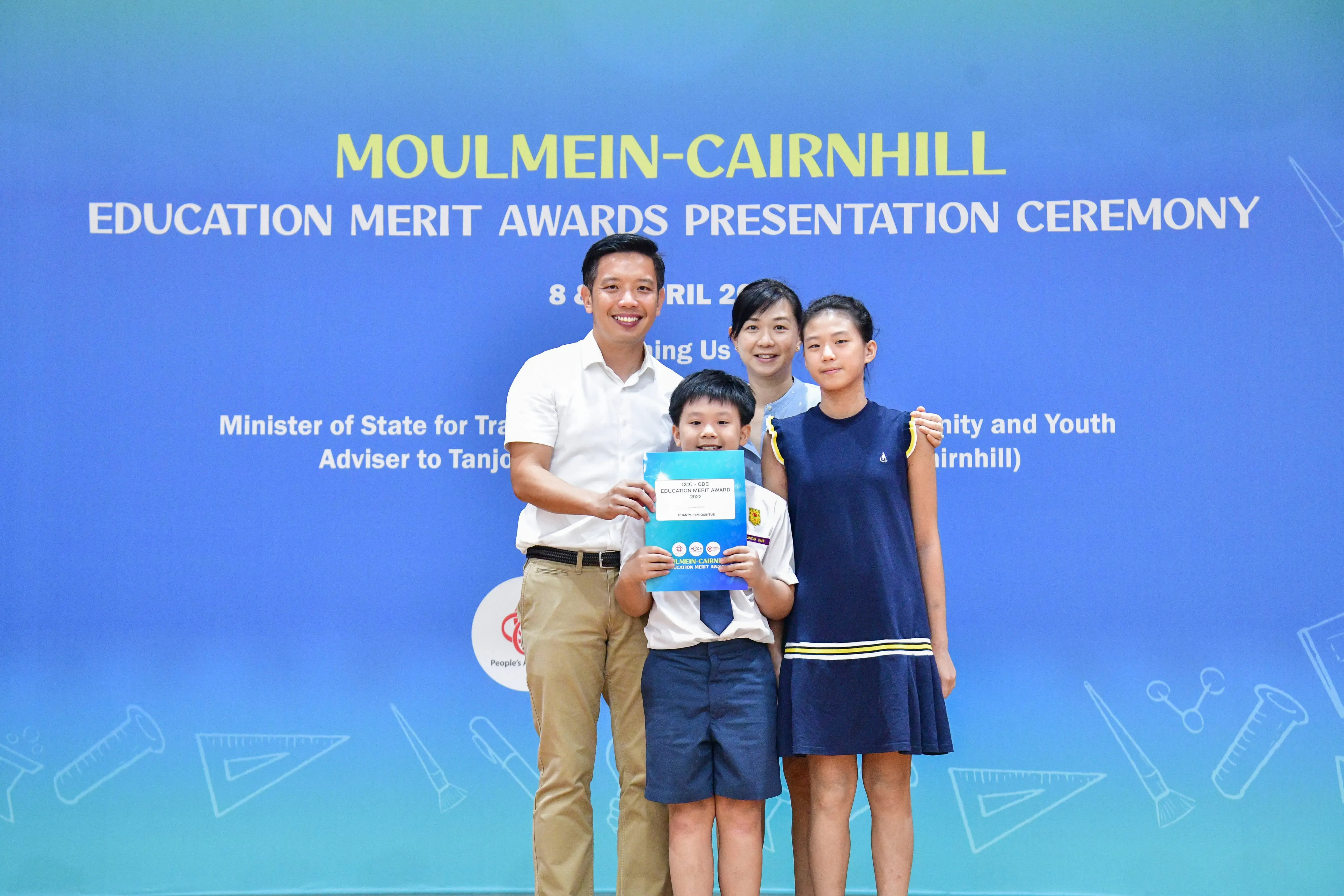 Four people pose at the Moulmein-Cairnhill Education Merit Awards Presentation Ceremony, with a boy holding a blue award folder.