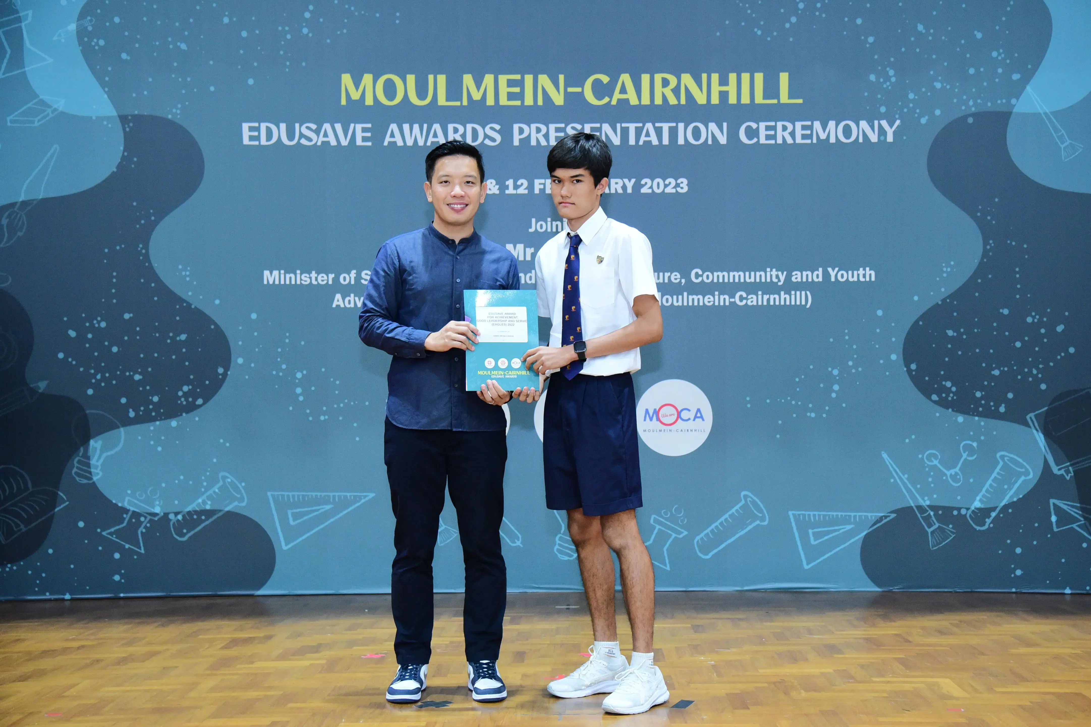 Two men hold a teal booklet at the Moulmein-Cairnhill Edusave Awards Presentation Ceremony 2023.