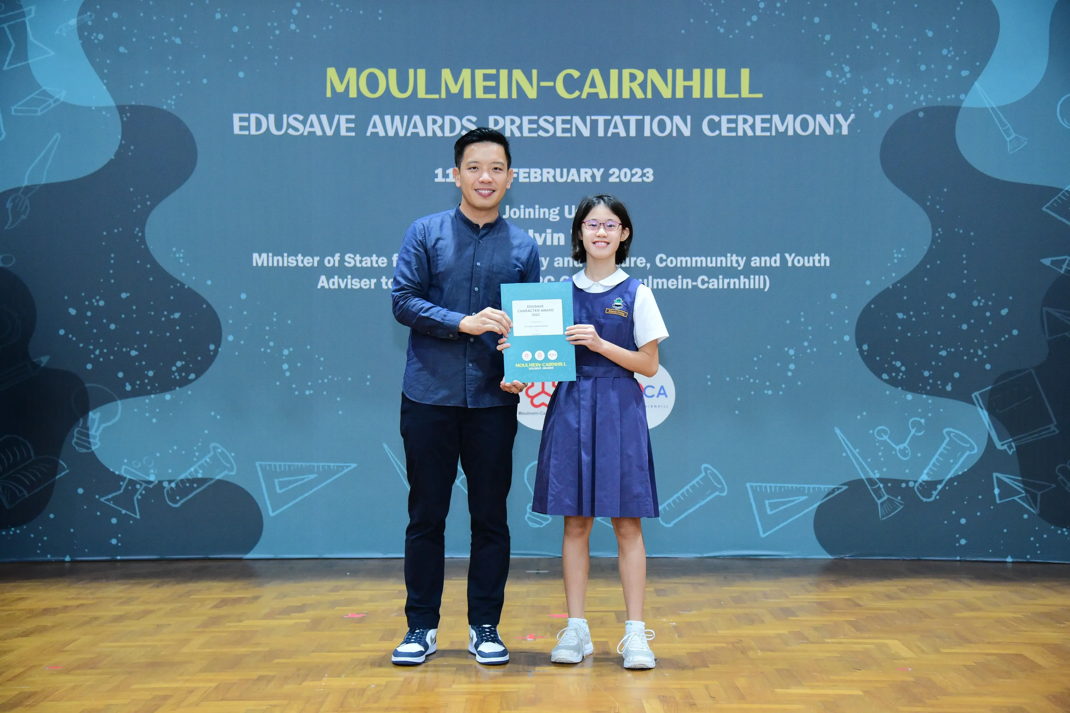 A man and a young girl in school uniform hold a certificate at the Moulmein-Cairnhill Edusave Awards Ceremony 2023.