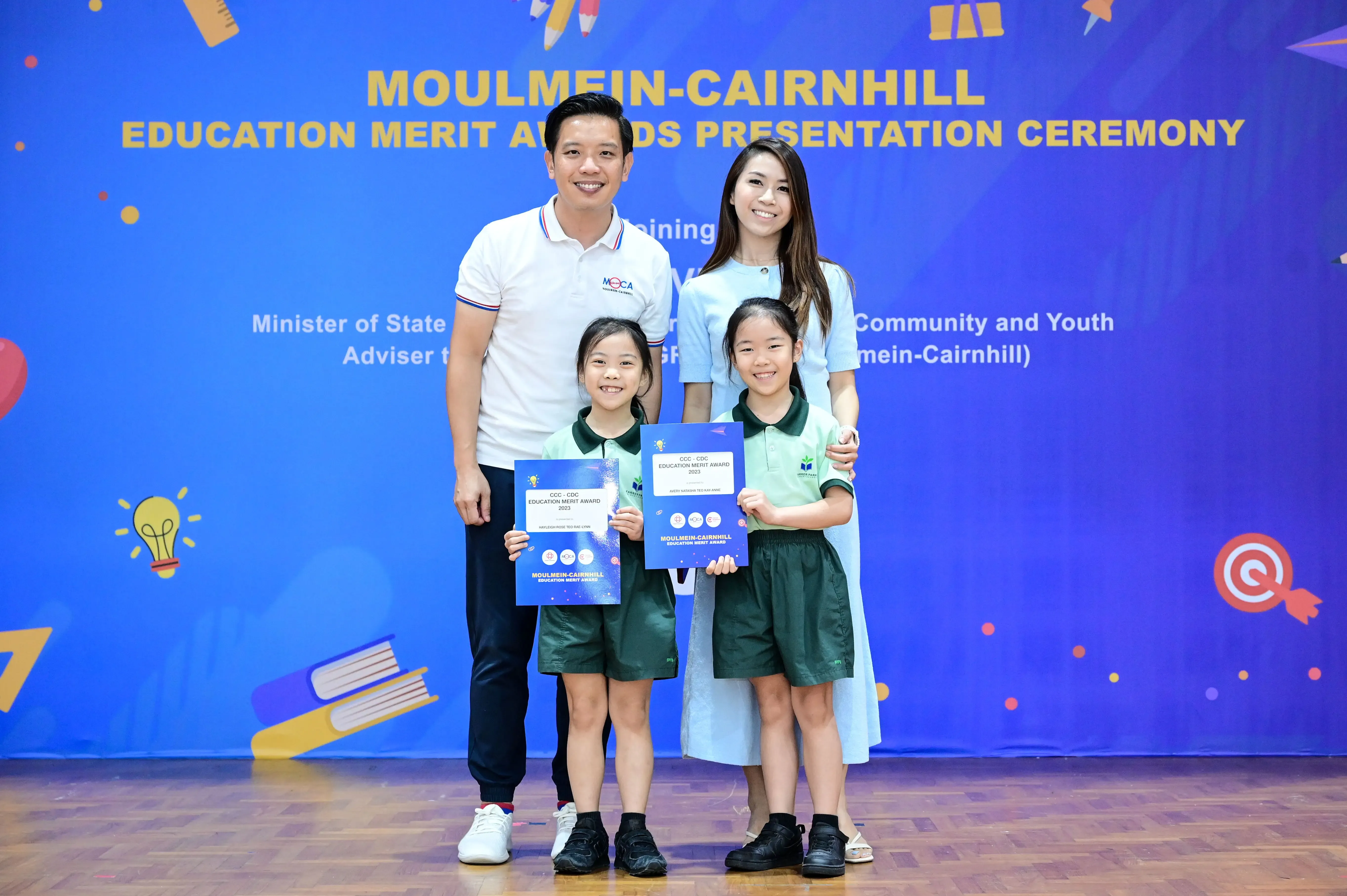 Two adults and two girls in school uniforms hold award certificates at the Moulmein-Cairnhill Education Merit Awards ceremony.