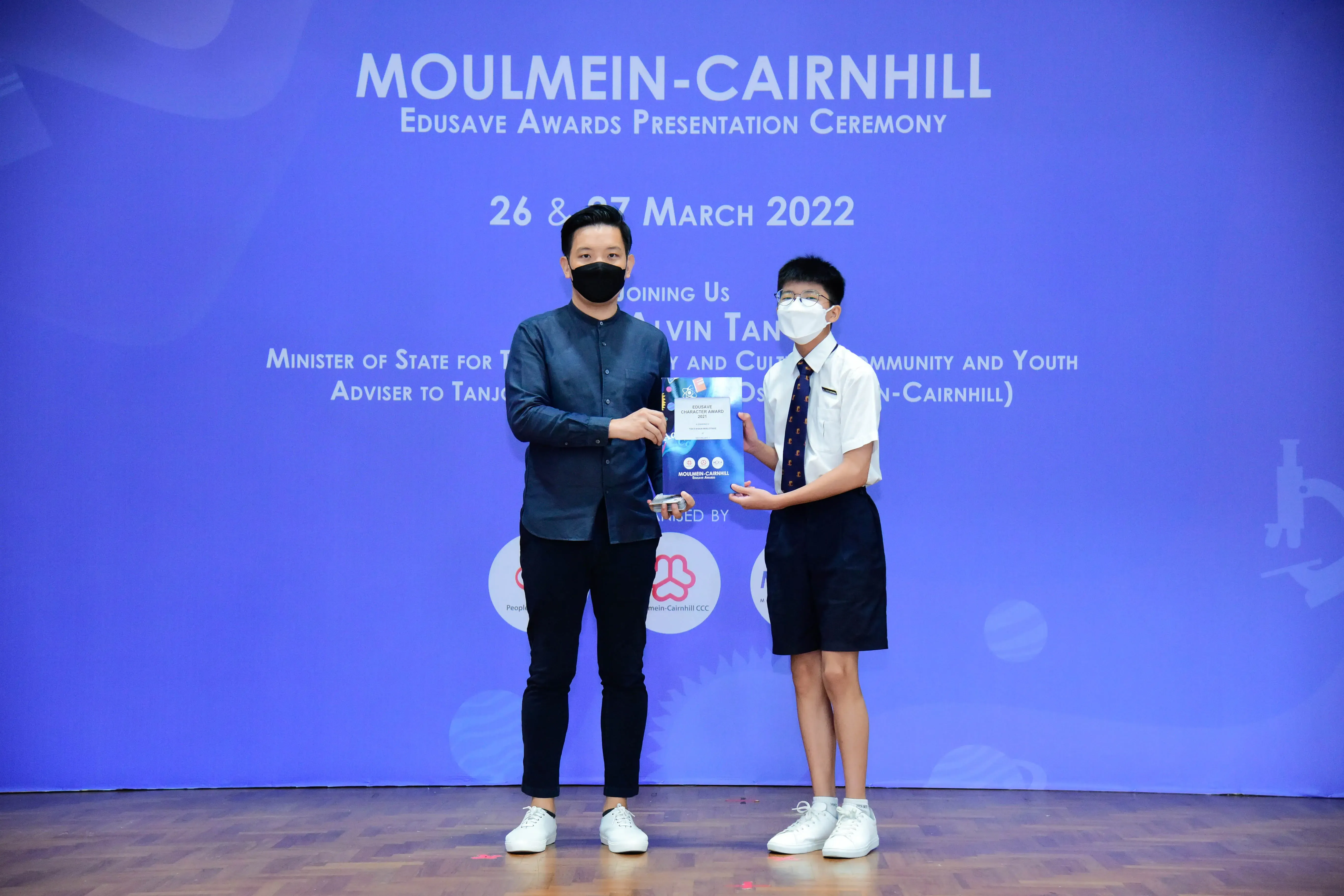 An adult and a uniformed student hold an award together on stage at the Moulmein-Cairnhill Edusave Awards Ceremony 2022.