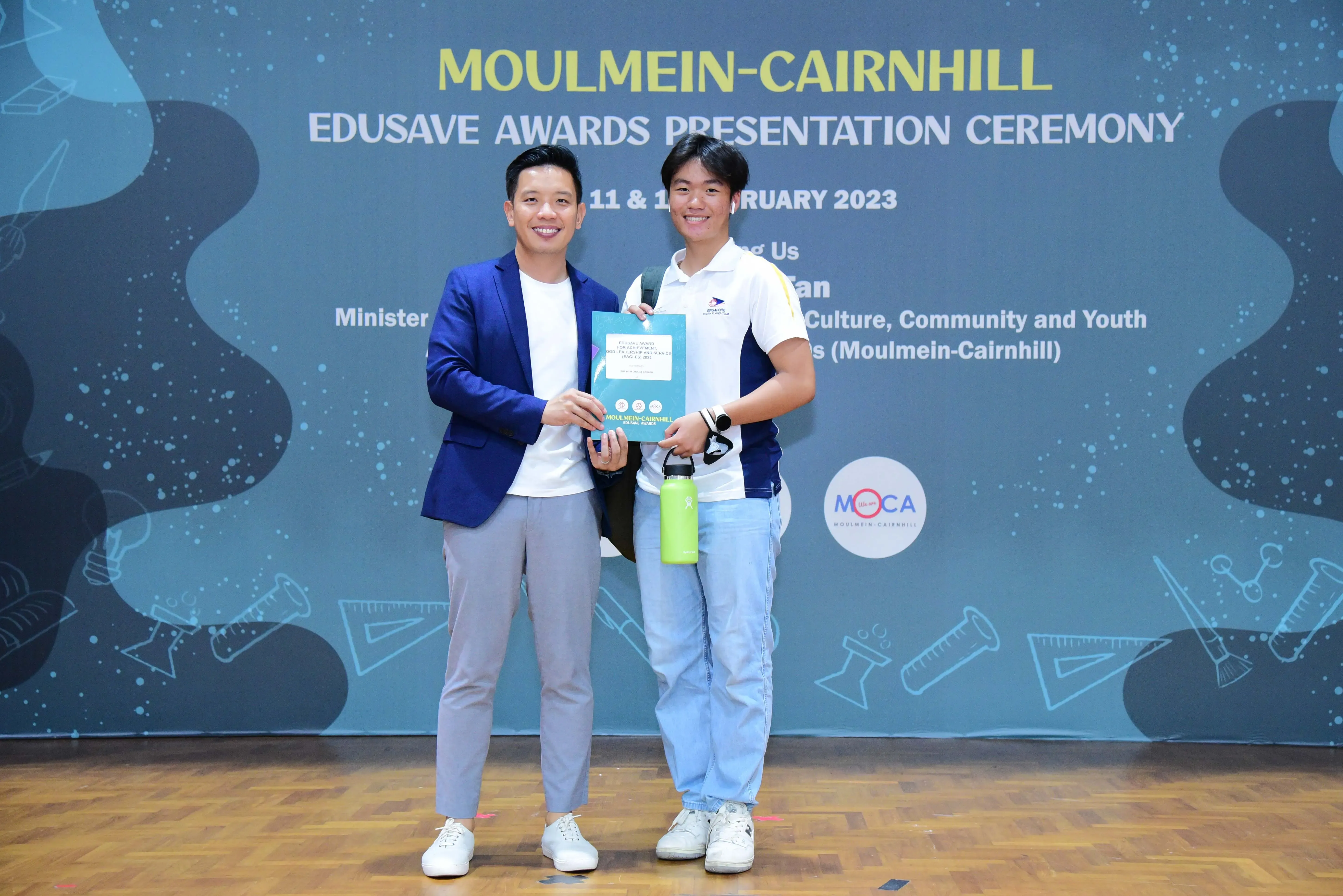 Two men smile holding a booklet at the Moulmein-Cairnhill Edusave Awards Presentation Ceremony 2023.