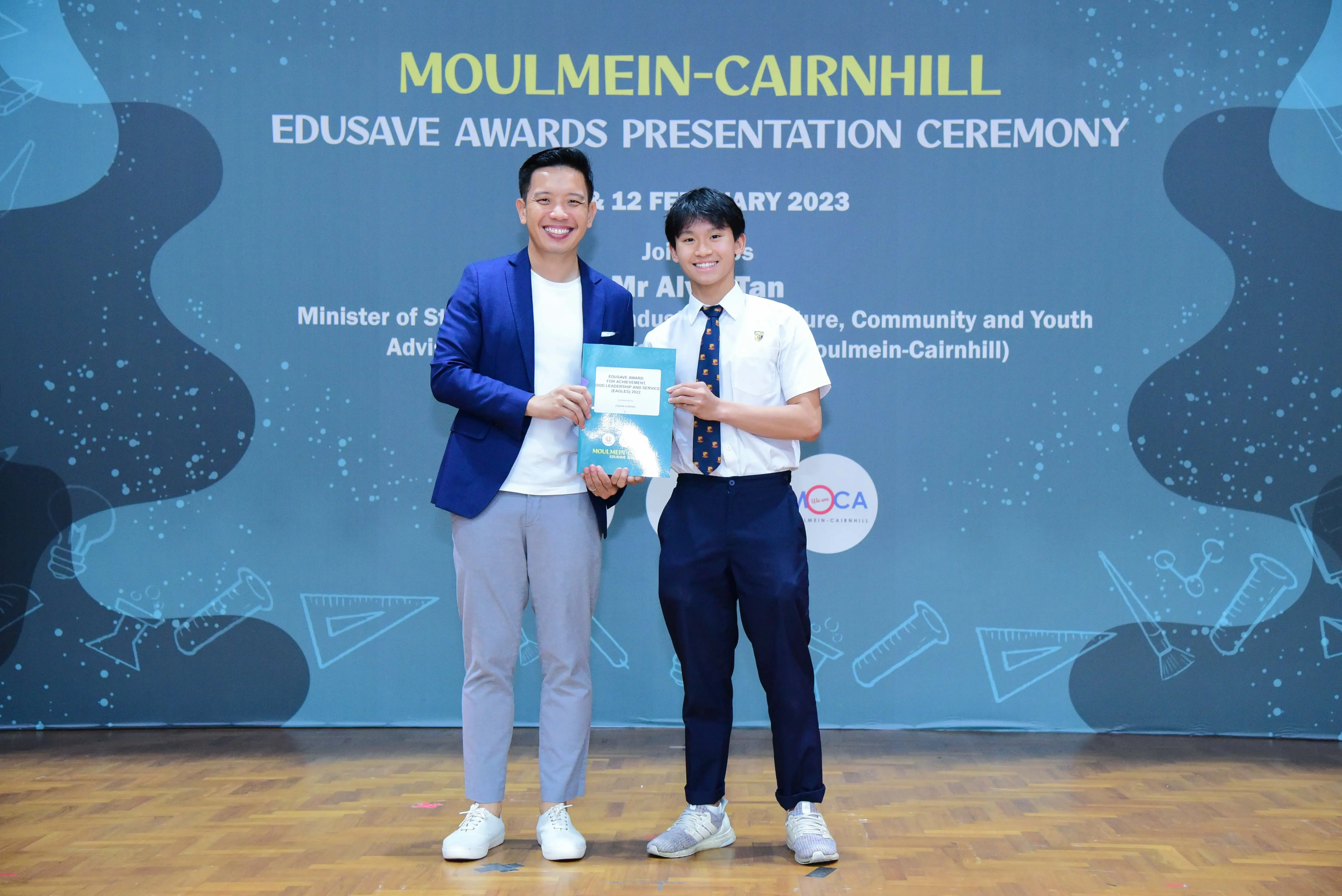 Two smiling males hold a booklet at the Moulmein-Cairnhill Edusave Awards Presentation Ceremony on 12 February 2023.