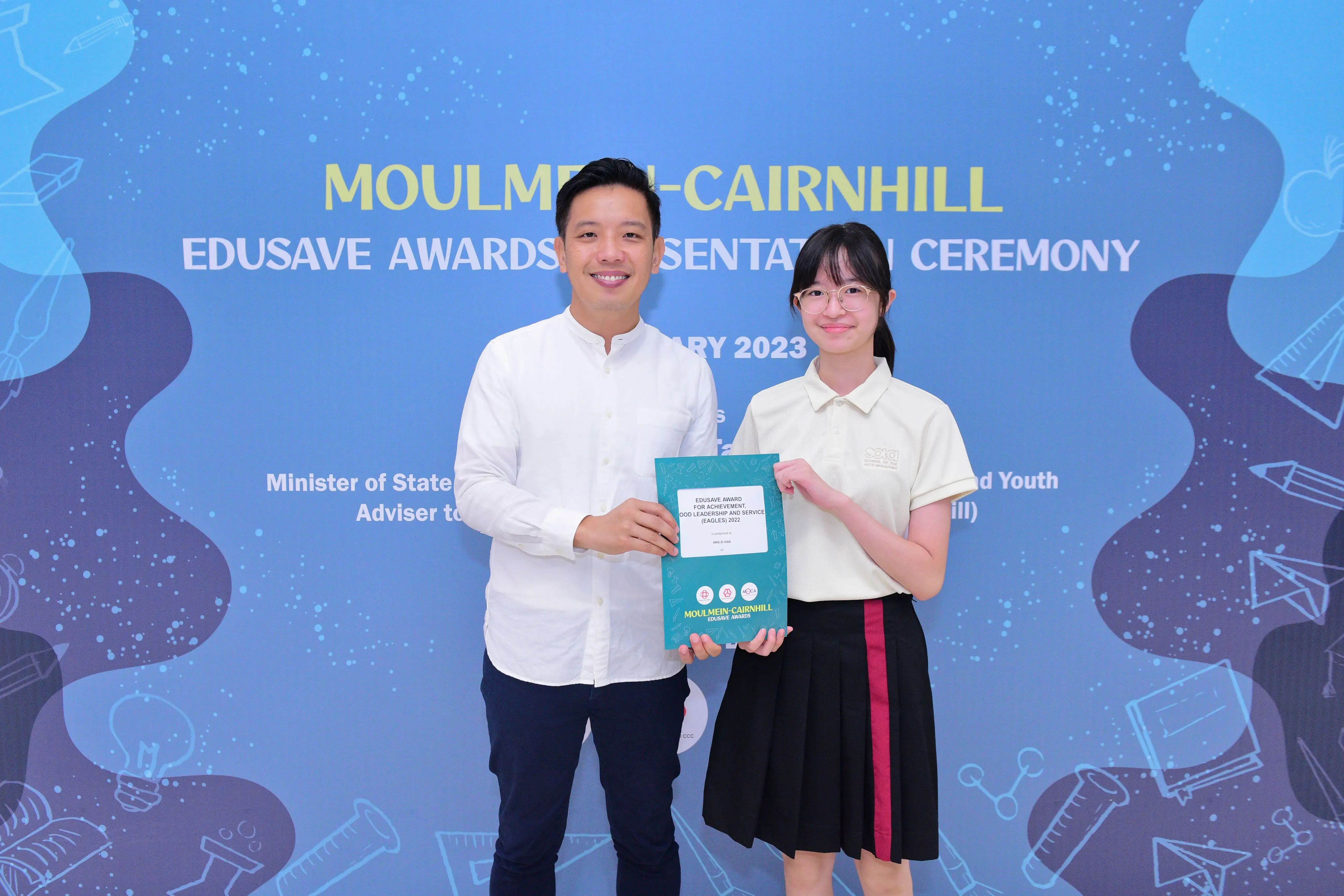 A man and a young girl hold a teal award booklet at the Moulmein-Cairnhill Edusave Awards Ceremony 2023.