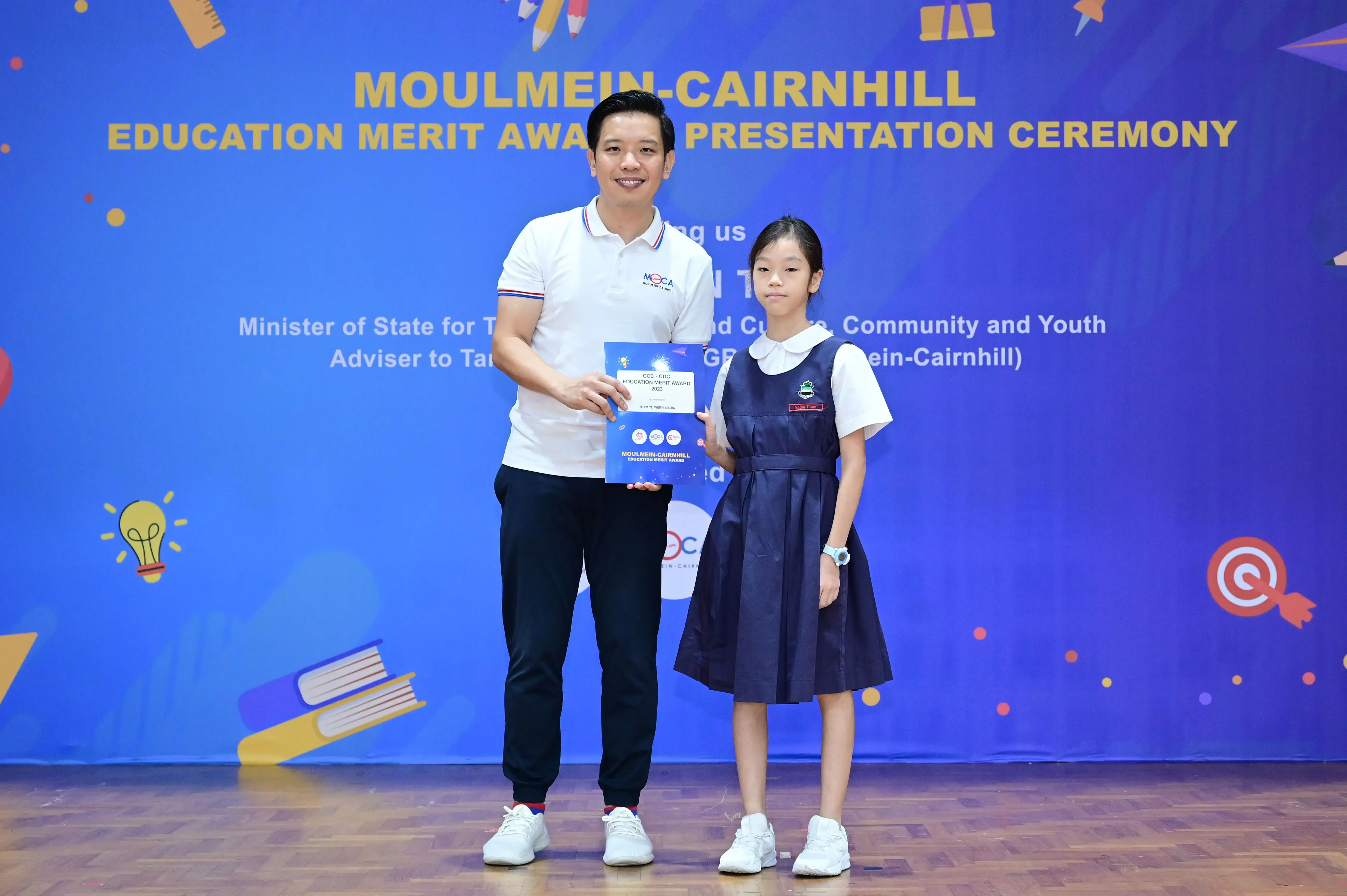 A man and a young girl in school uniform hold an award certificate at the Moulmein-Cairnhill Education Merit Award ceremony.
