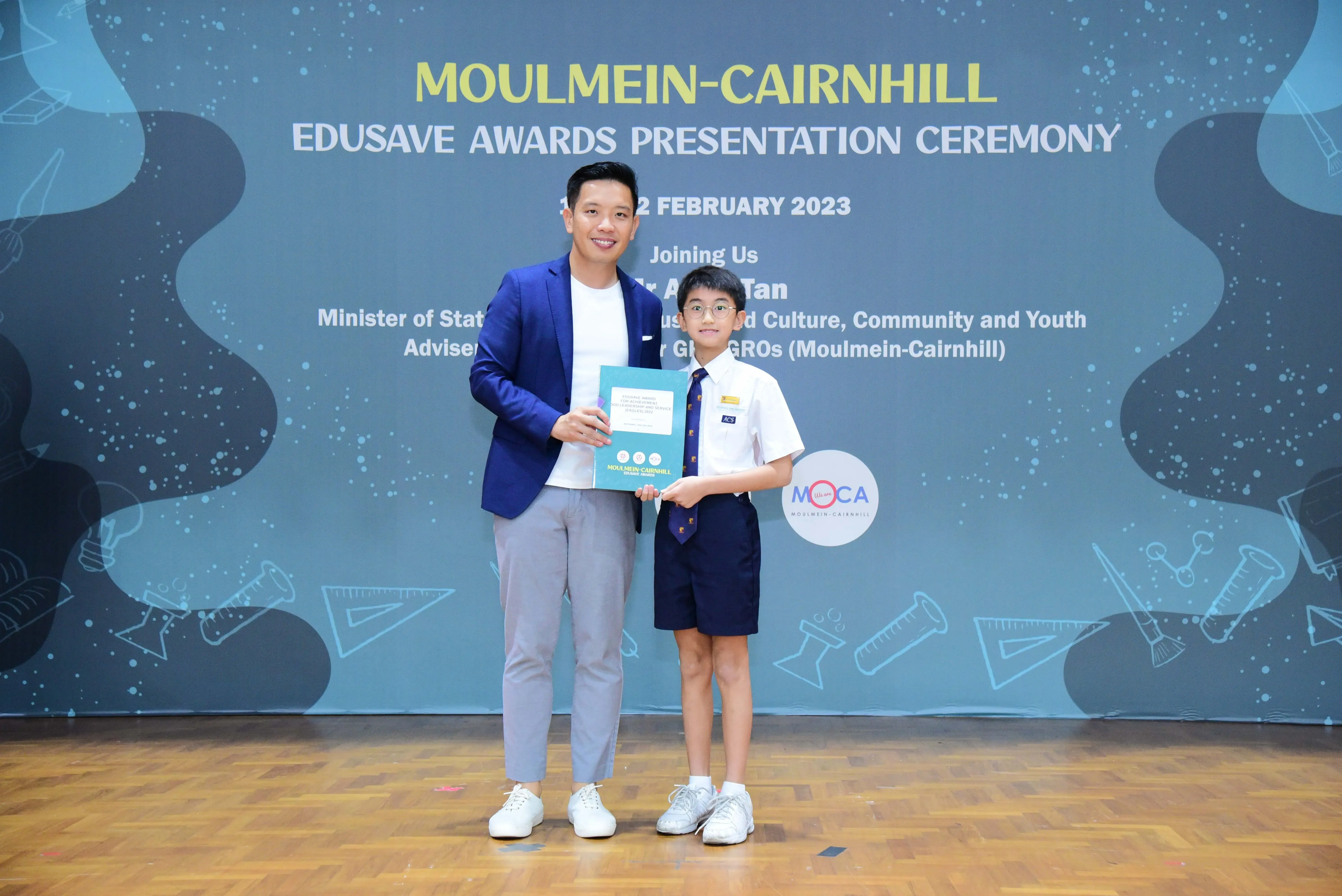 A man in a blue blazer and a young boy in school uniform hold a certificate at the Moulmein-Cairnhill Edusave Awards 2023.