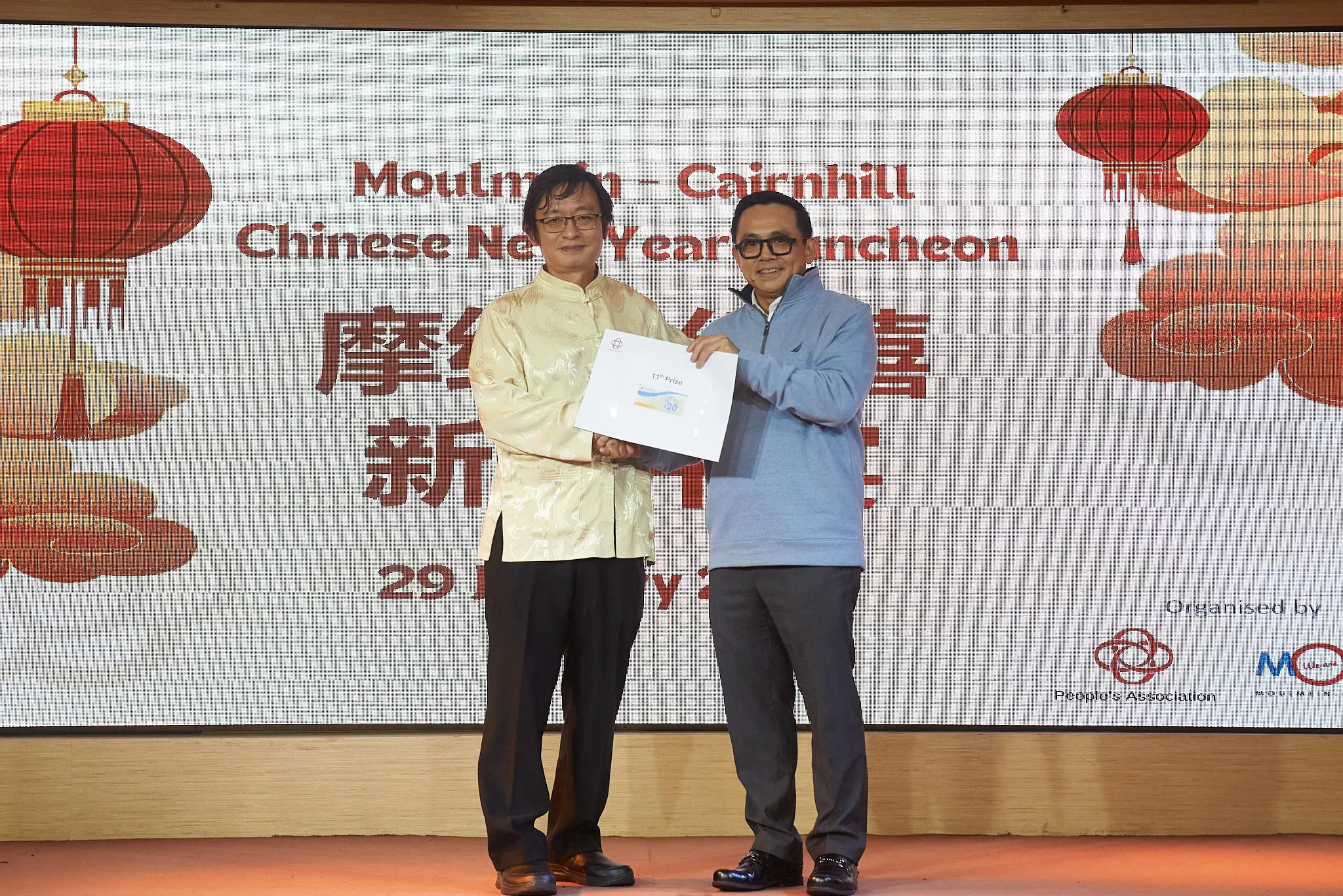 Two men hold an 11th prize envelope on stage at the Moulmein-Cairnhill Chinese New Year Luncheon.