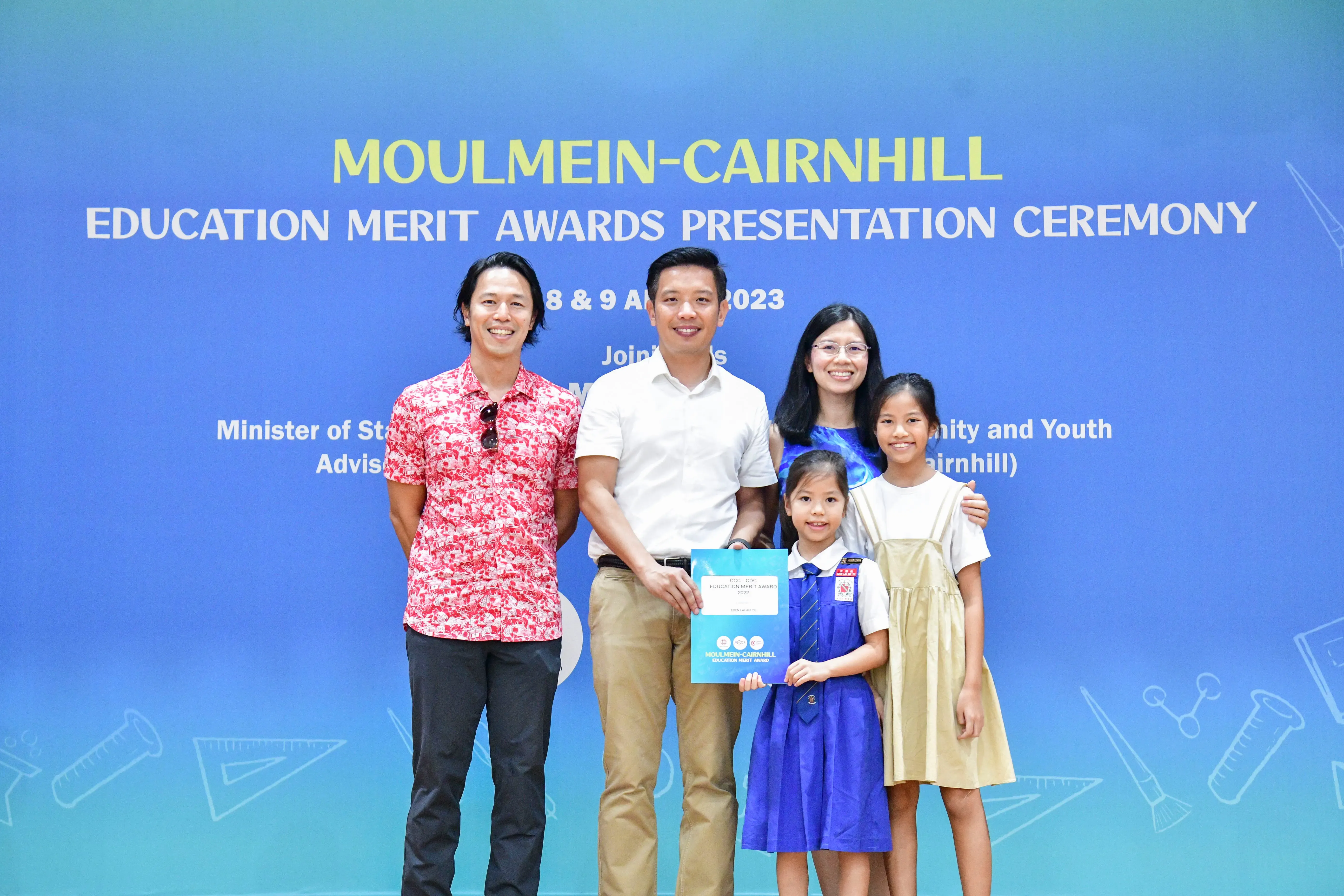 Five people pose before a blue Moulmein-Cairnhill Education Merit Awards Presentation Ceremony banner in 2023.