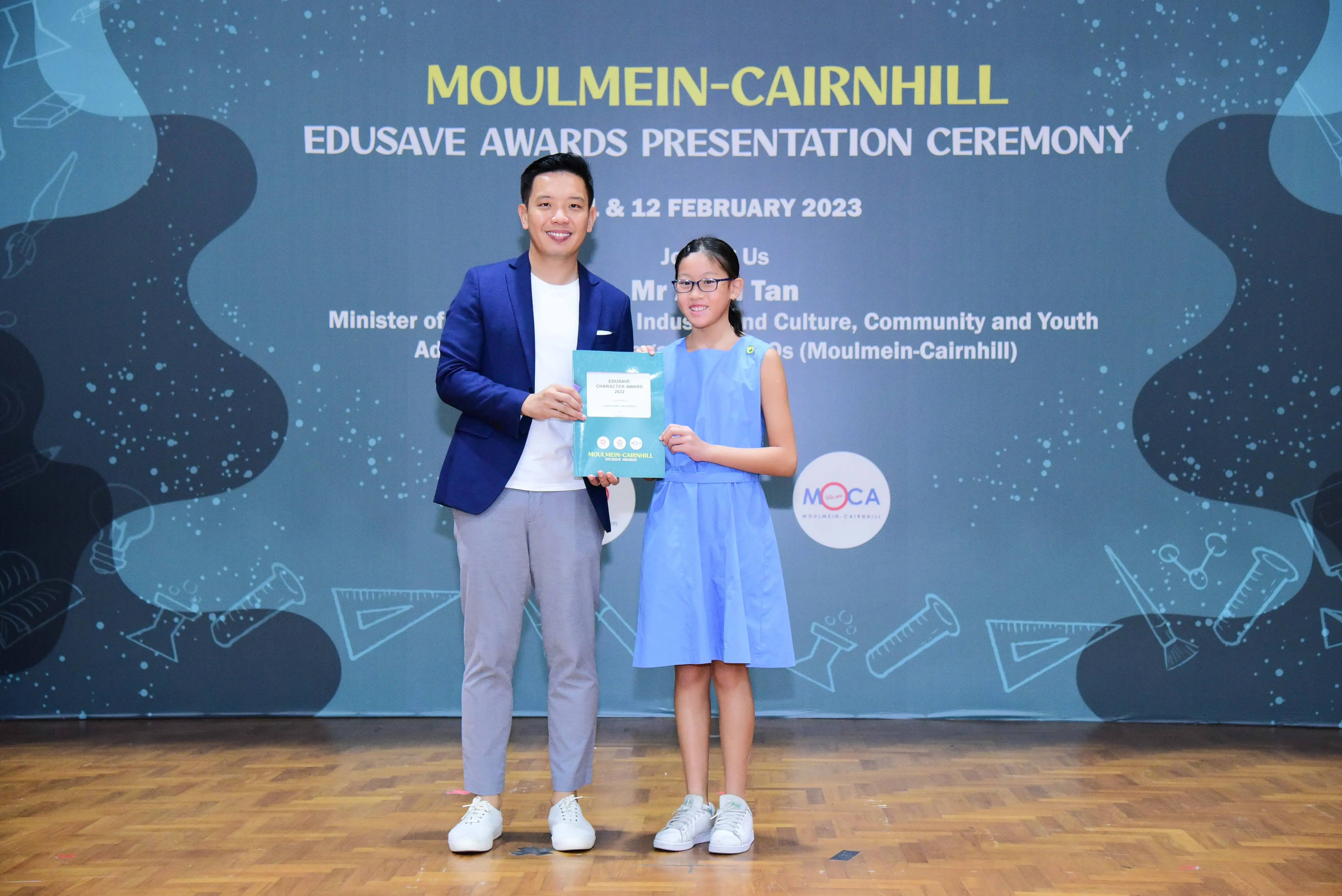 A man in a blue blazer and a young girl in a blue dress hold a certificate at the Moulmein-Cairnhill Edusave Awards 2023.