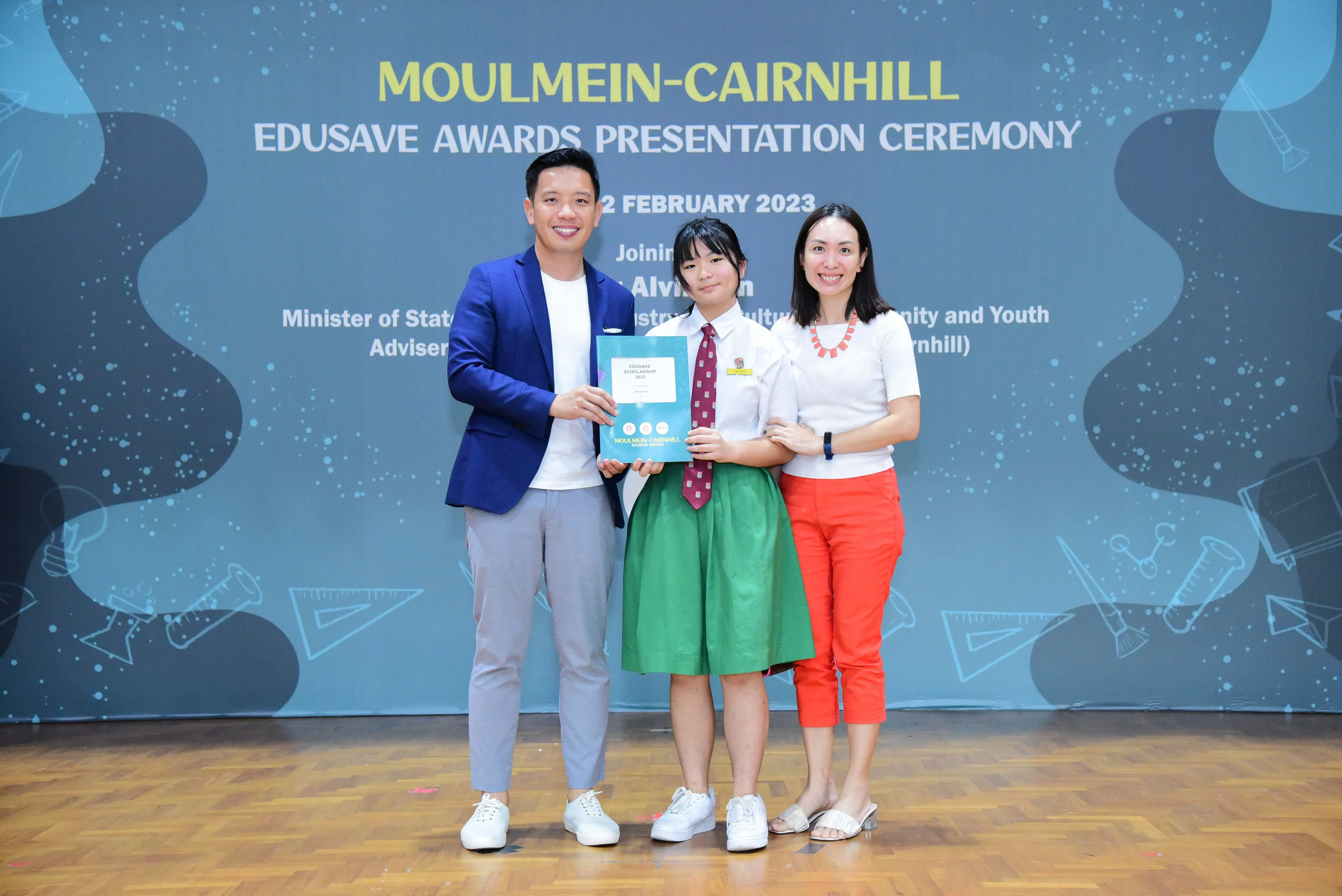 Three people pose on a stage at the Moulmein-Cairnhill Edusave Awards Presentation Ceremony, February 2023.