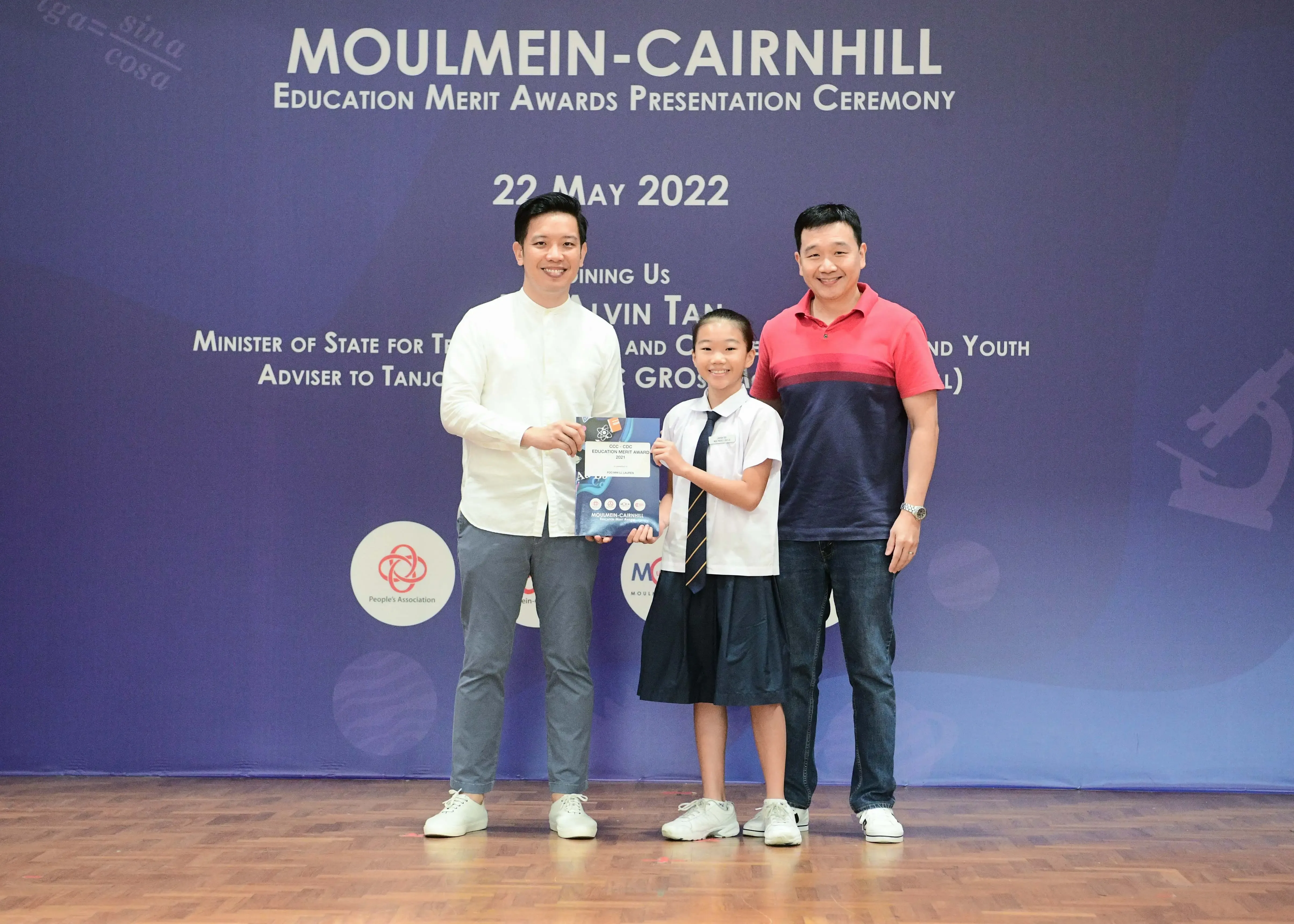 Two men and a uniformed student hold an award at the Moulmein-Cairnhill Education Merit Awards Ceremony, May 2022.