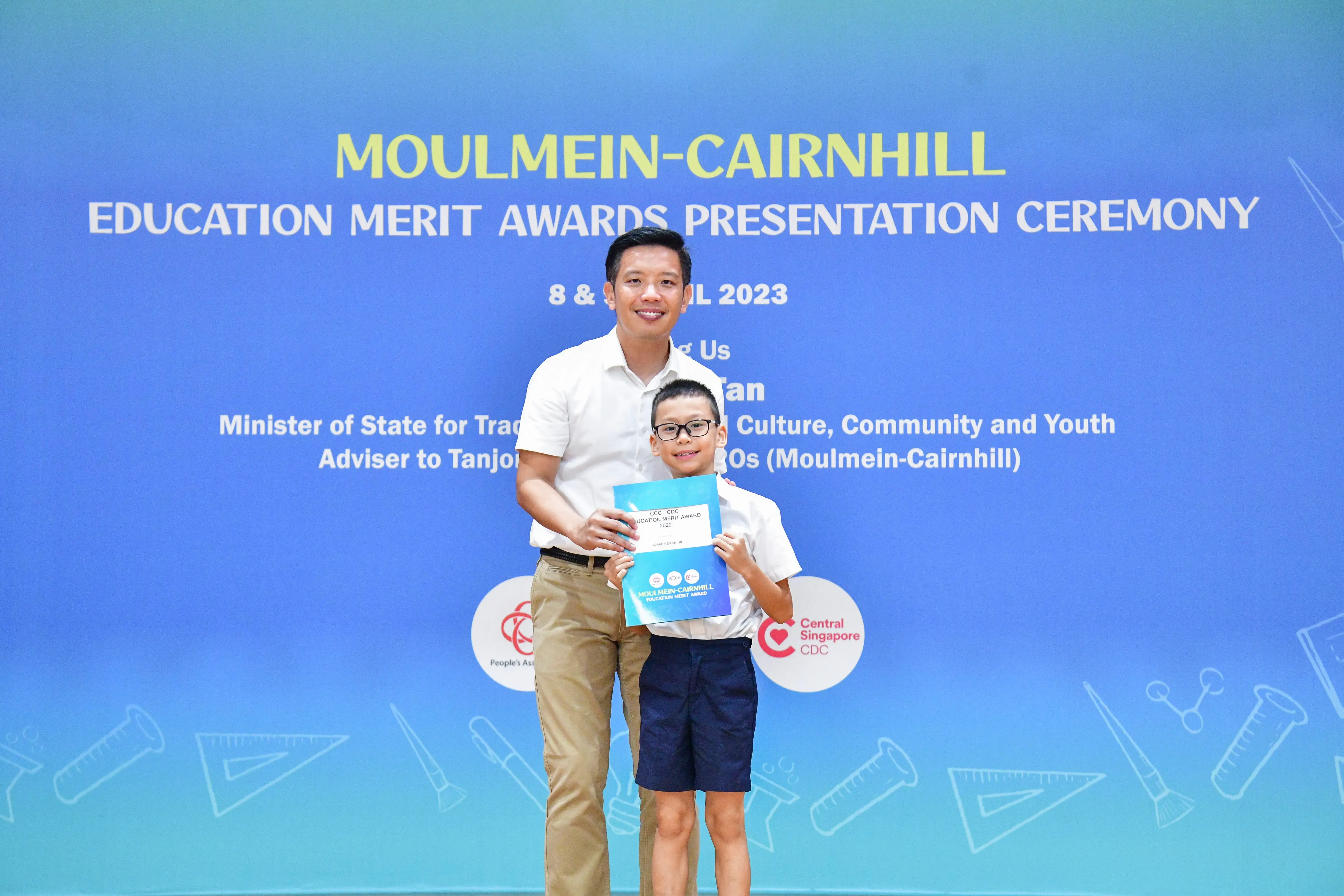 A man and a young boy holding a blue award certificate stand before the Moulmein-Cairnhill Education Merit Awards banner.