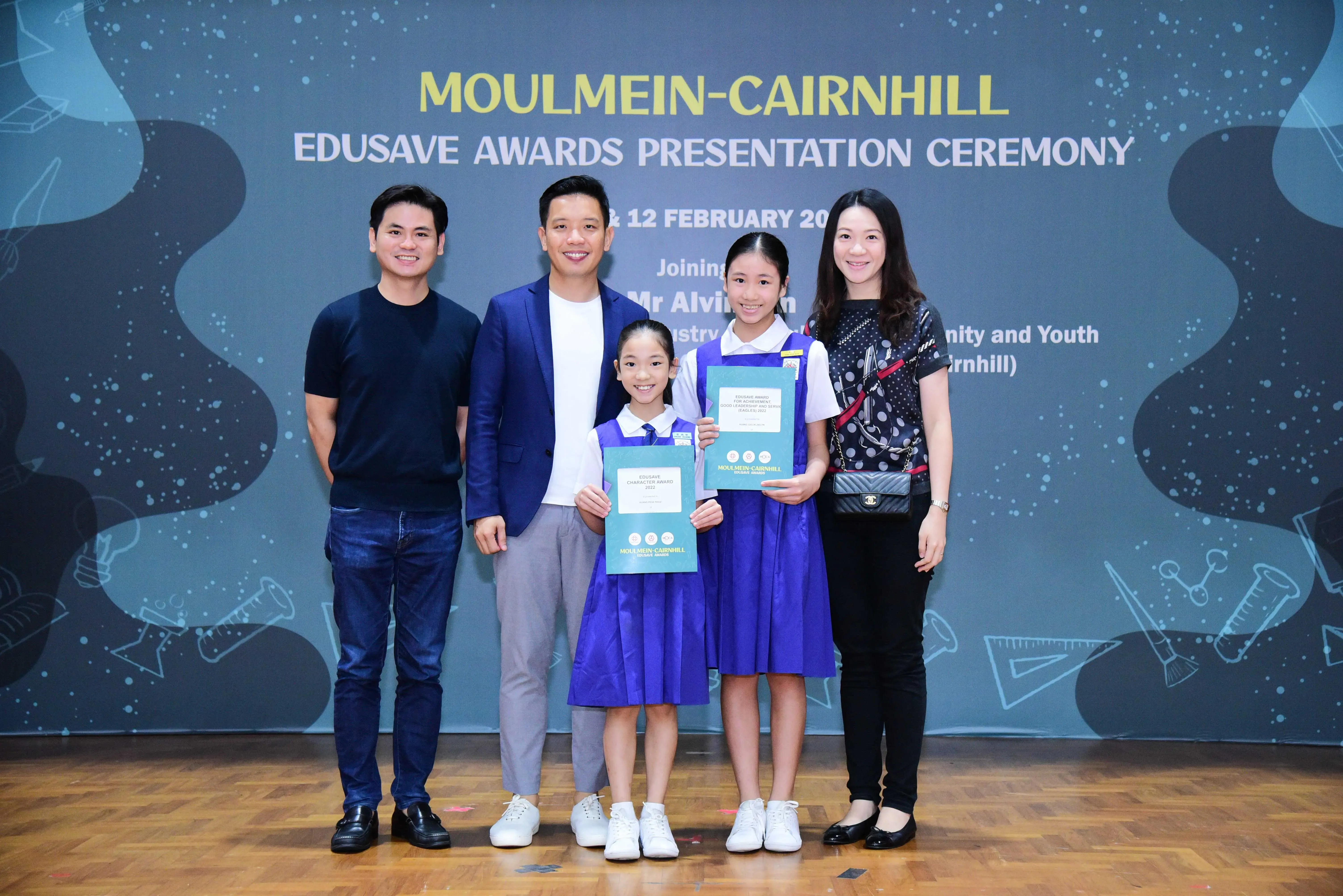 Five people pose at the Moulmein-Cairnhill Edusave Awards Presentation Ceremony, two girls in blue uniforms holding certificates.