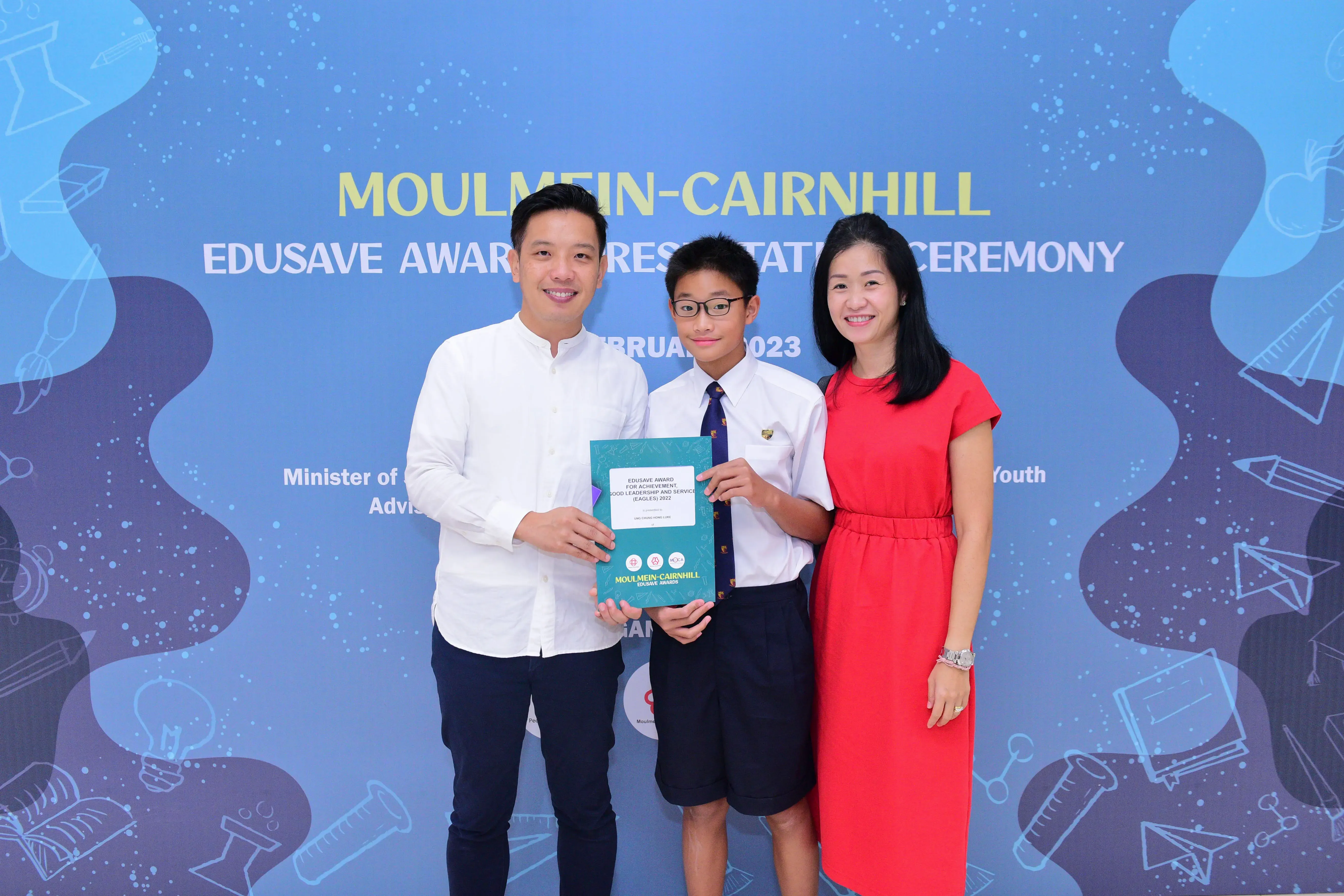 A boy in school uniform holds a certificate flanked by two smiling adults at the Moulmein-Cairnhill Edusave Awards Ceremony 2023.