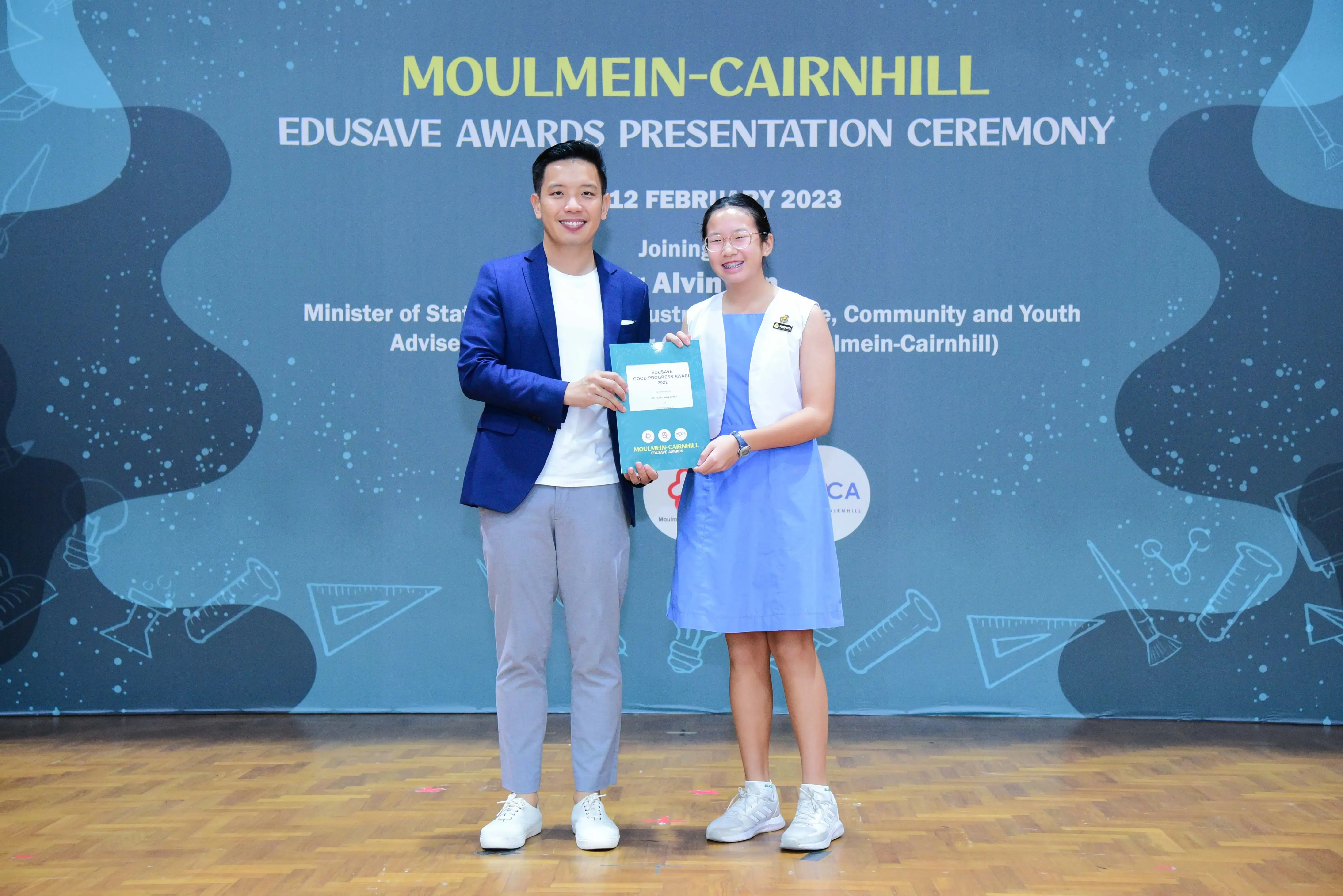 A man in a blue blazer and a young girl in a blue dress hold an award at the Moulmein-Cairnhill Edusave Awards Ceremony.