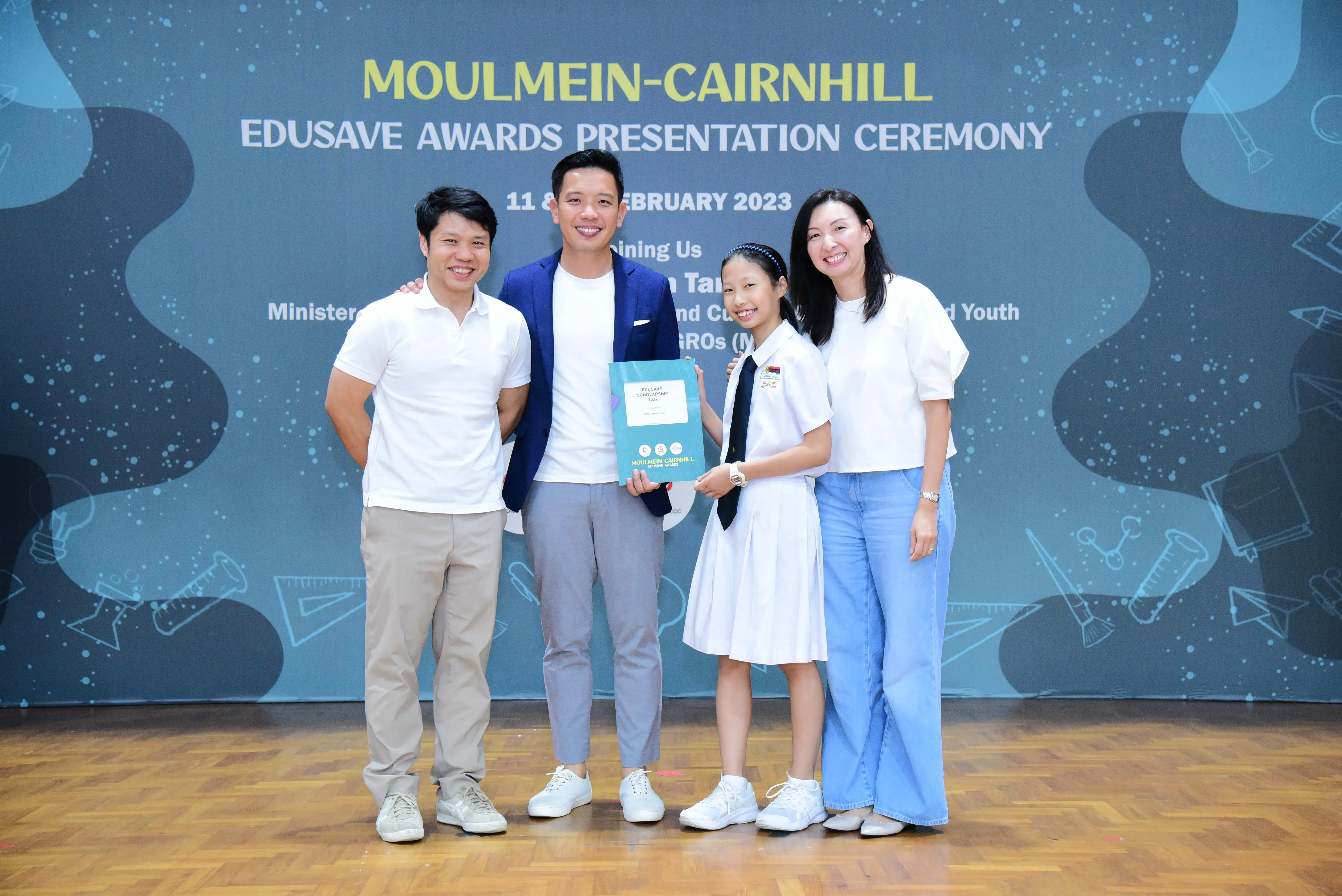 Four people pose smiling at the Moulmein-Cairnhill Edusave Awards Presentation Ceremony, with a student holding an award certificate.