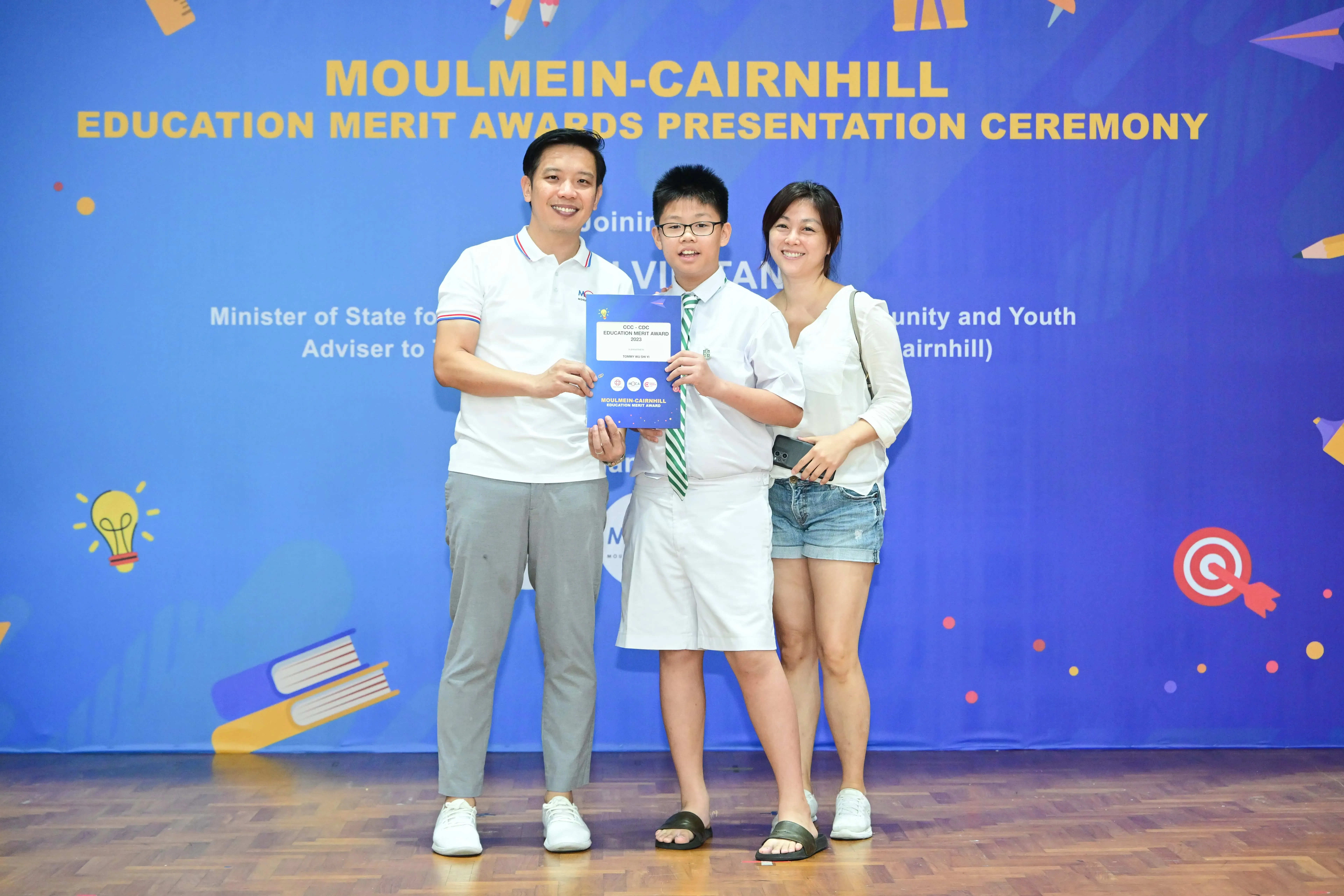 A boy in school uniform holds an award certificate, flanked by two smiling adults, at the Moulmein-Cairnhill Education Merit Awards ceremony.