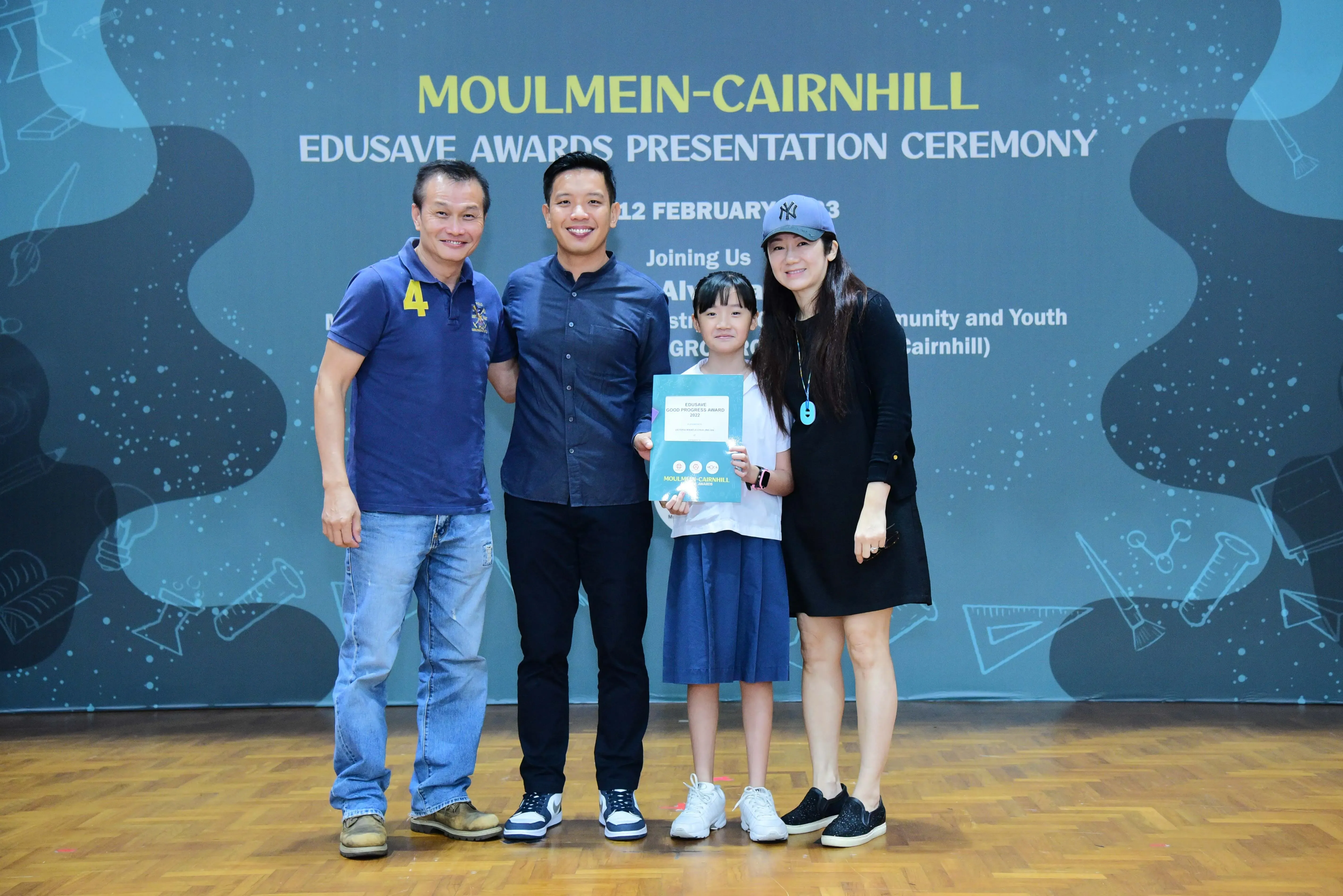Four people pose at the Moulmein-Cairnhill Edusave Awards Presentation Ceremony, with a young girl holding a certificate.
